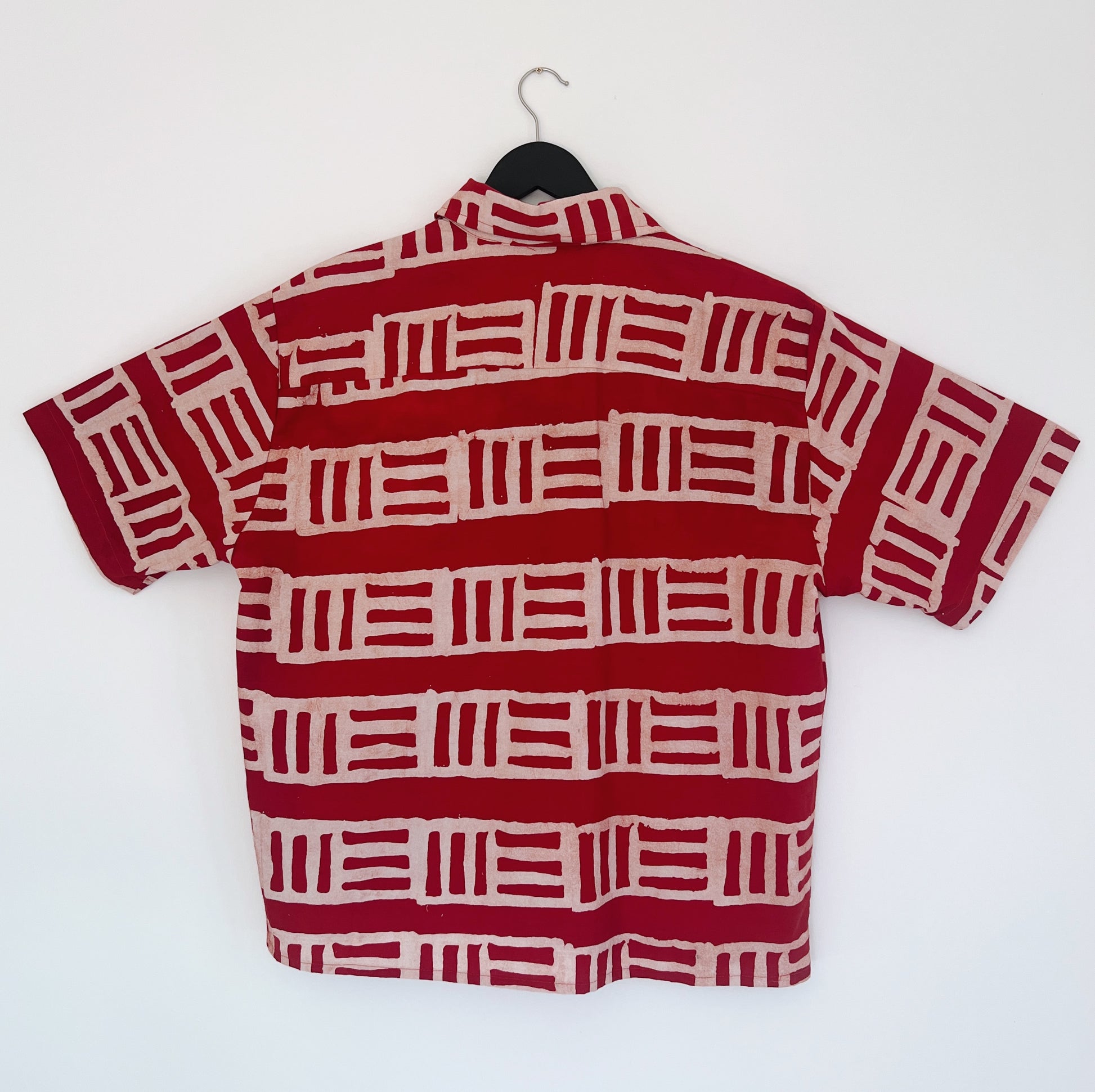 Back view of the Sudi Shirt in Brick, hanging on a black hanger against a white wall, showing the relaxed boxy silhouette, short sleeves, and bold hand-printed brick red and blush geometric block stripe pattern on 100% cotton Batiki fabric

