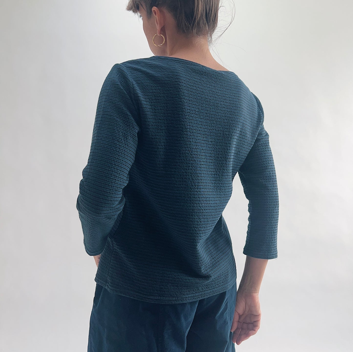 Back view of model wearing a deep teal tonal textured stripe boatneck tee with 3/4 sleeves, showing the wide boatneck and curved hem, styled with matching teal wide-leg pants.
