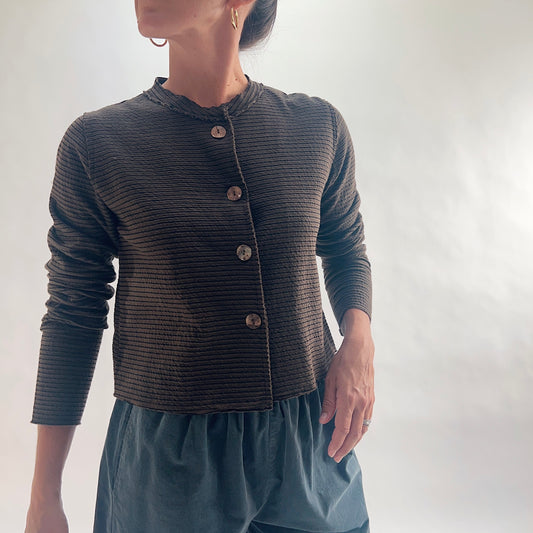 Close front view of model wearing a chocolate and black fine tonal stripe cropped cardigan with asymmetrical button closure, band neckline, and long sleeves, hand at side, showing the crinkle-texture fabric and cropped length, styled with teal wide-leg pants against a white background.
