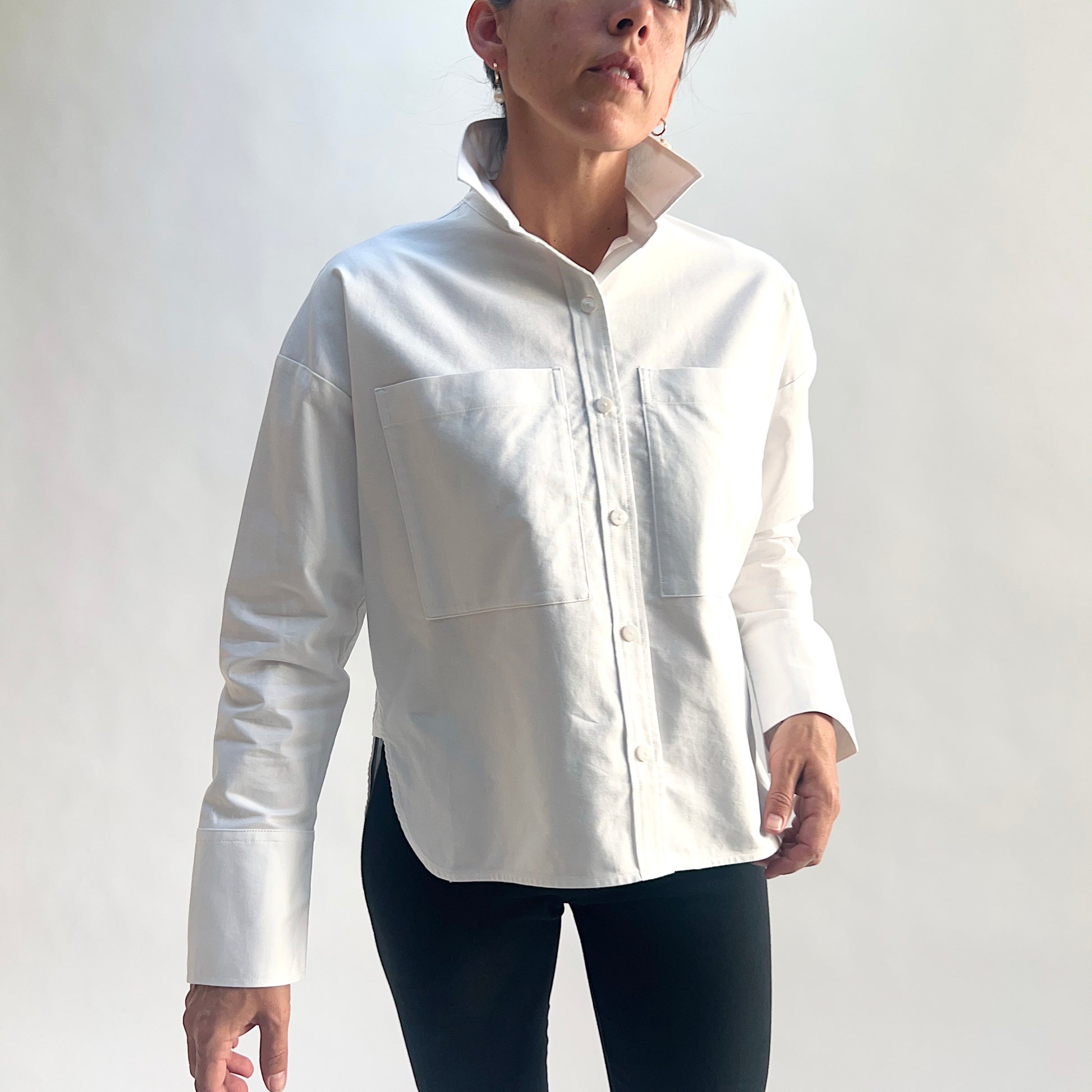 Full front view of model wearing a white cotton oxford boyfriend shirt, arms at sides, showing the point collar, two large chest patch pockets, full button front, and wide barrel cuffs, styled with black slim pants against a white background.
