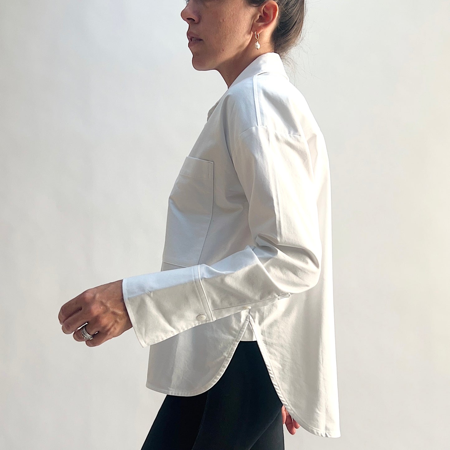 Side view of model wearing a white cotton oxford boyfriend shirt in motion, showing the relaxed oversized silhouette, longer curved shirttail, side hem slit, wide barrel cuff, and chest patch pocket, styled with black slim pants against a white background.
