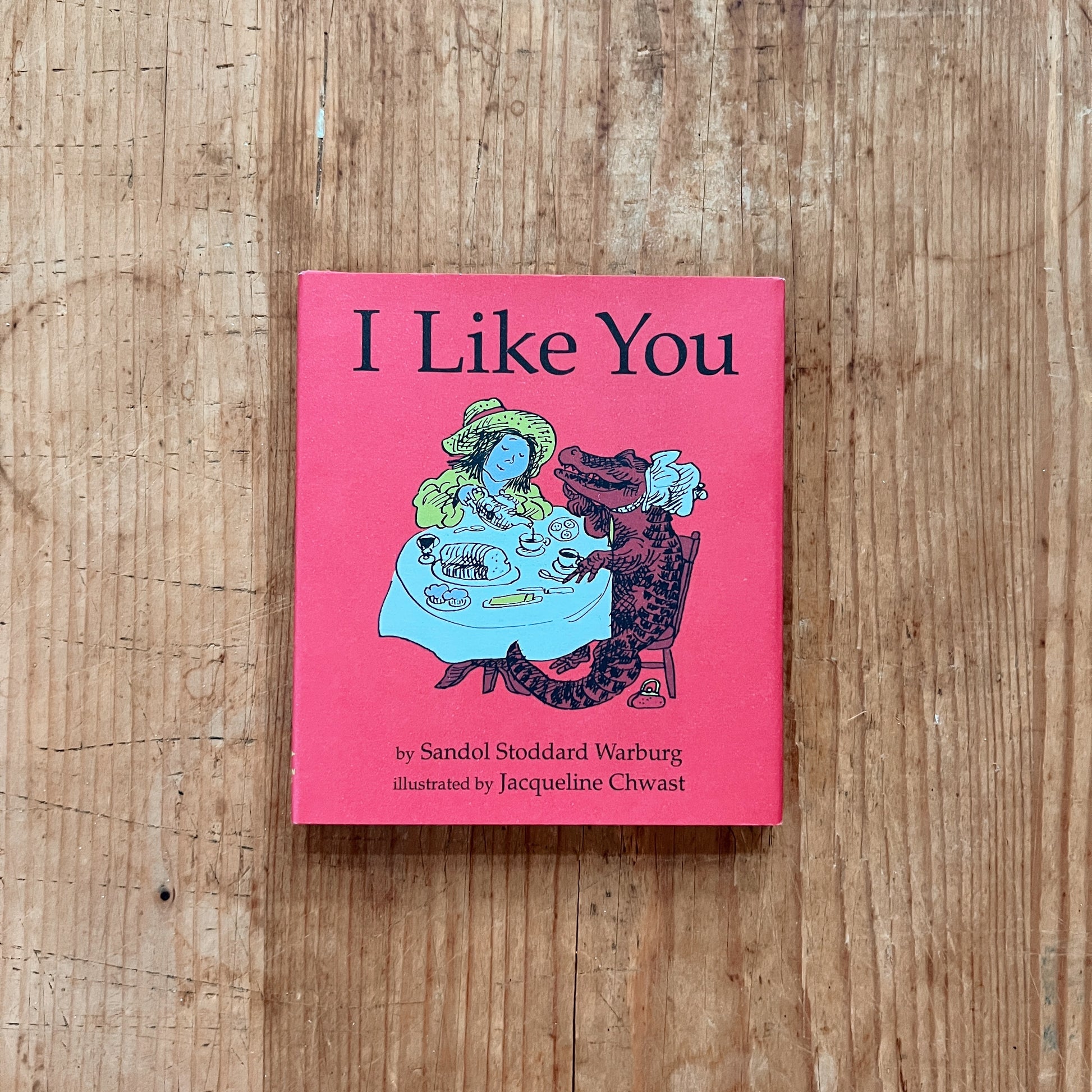 Front cover of I Like You by Sandol Stoddard Warburg, featuring a vivid red background with Jacqueline Chwast's black ink and color illustration of a young girl in a yellow-green dress and wide-brimmed hat sitting across a blue-clothed tea table from a large brown crocodile wearing a pearl necklace and bib, with teacups, a cake, cookies, and a bread loaf on the table between them. The title is set in large black serif lettering at the top, and the author and illustrator credits appear at the bottom, display