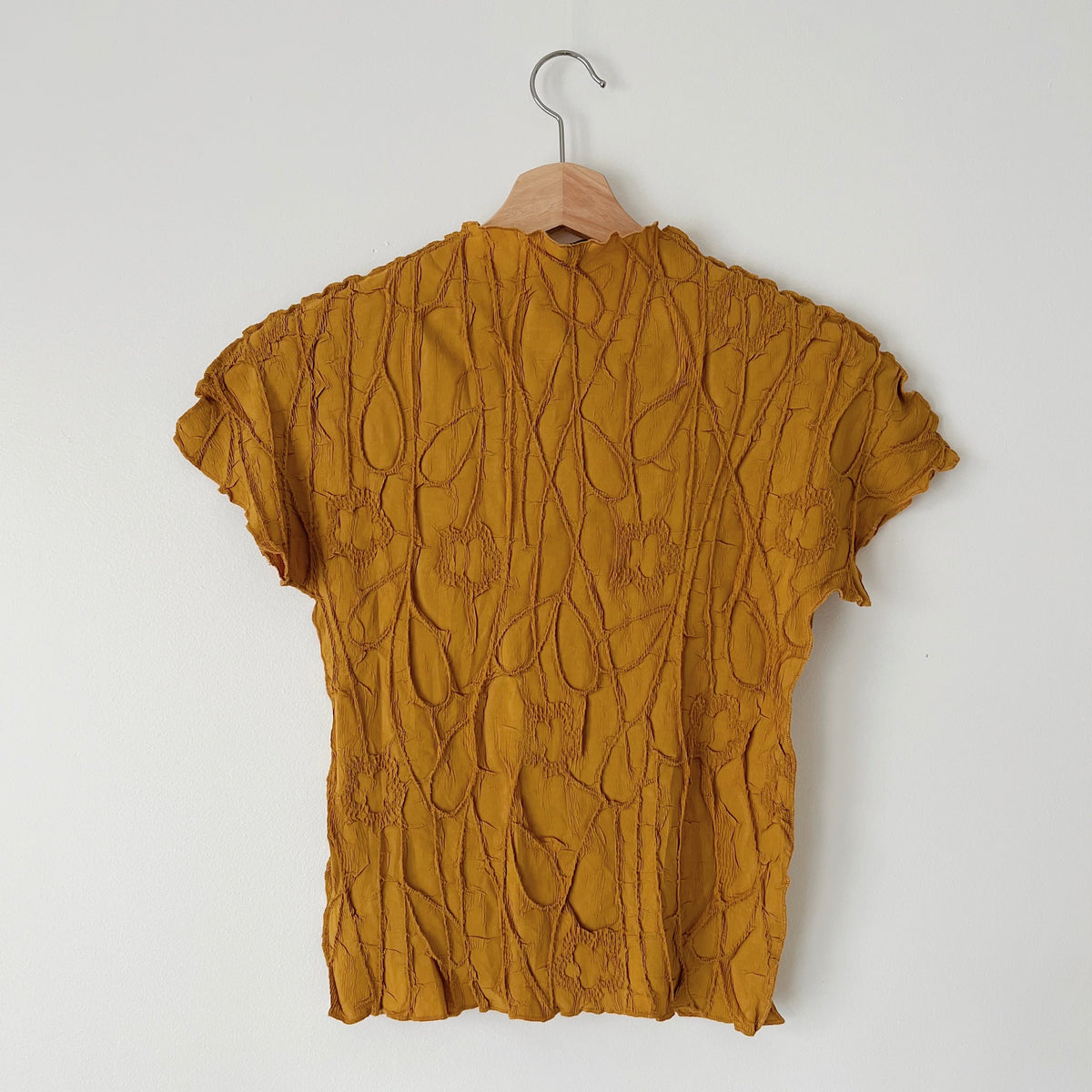 Back view of the mustard short sleeve crinkle top on a wooden hanger, showing its textured fabric.
