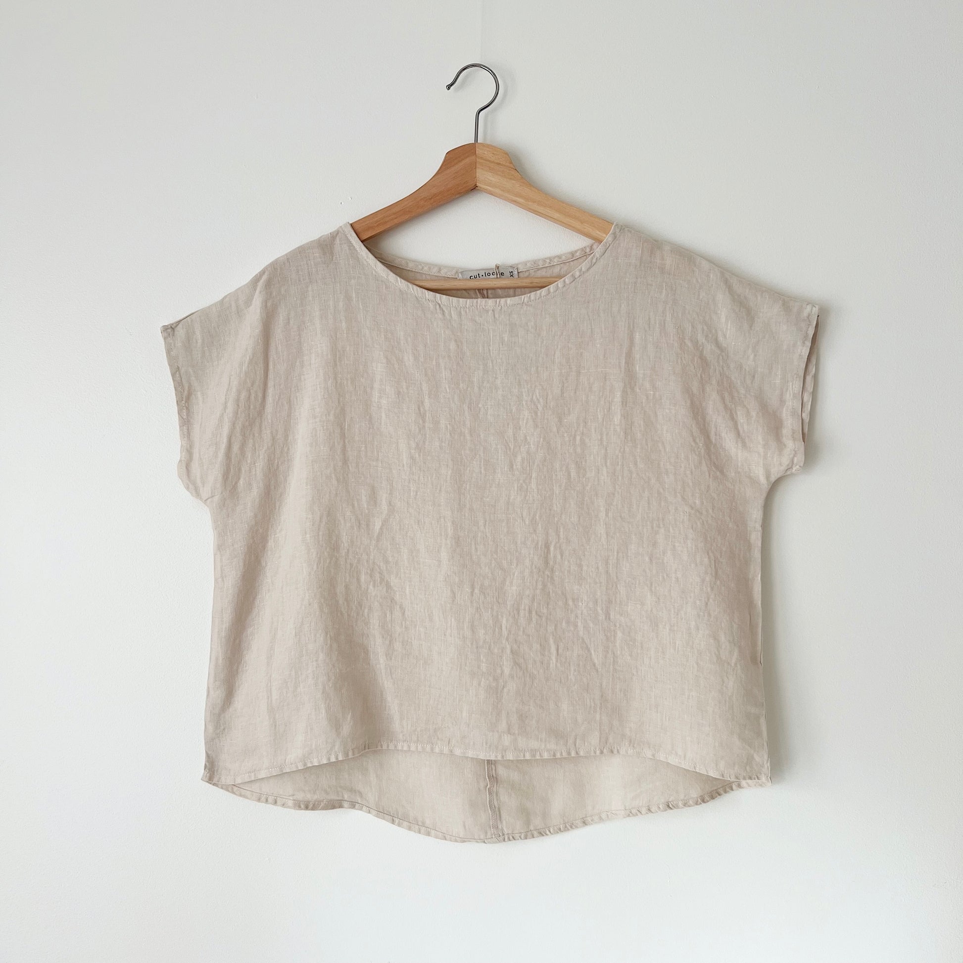 A beige high-low hem linen tee hanging on a wooden hanger against a white wall.