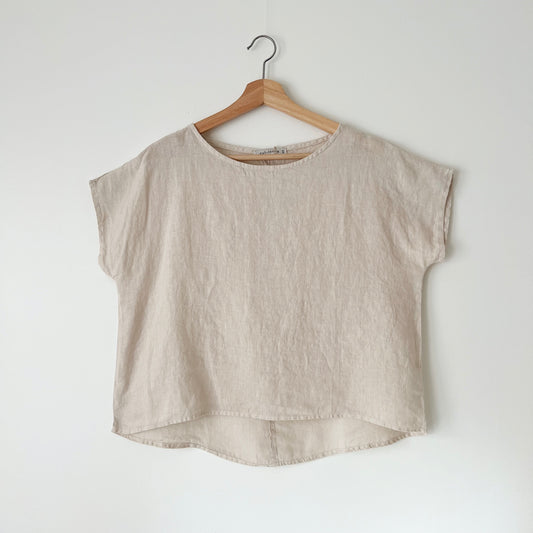 A beige high-low hem linen tee hanging on a wooden hanger against a white wall.