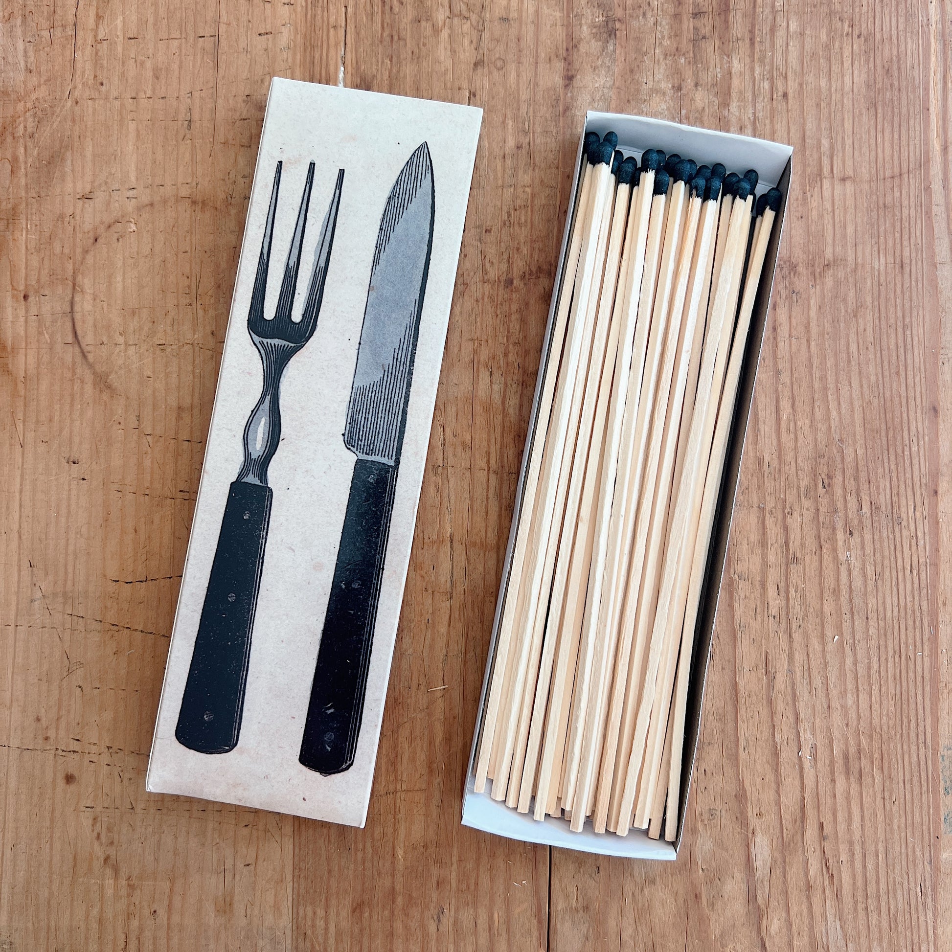 Dotter Silverware Matches box open on warm wood surface, top view showing kraft paper box lid with vintage engraving-style black illustration of a fork and carving knife side by side, with 50 long wooden matches with black tips fanned out in the open box tray
