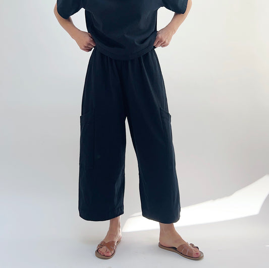 A person standing against a white background, wearing black cotton pasha pants and a black top.