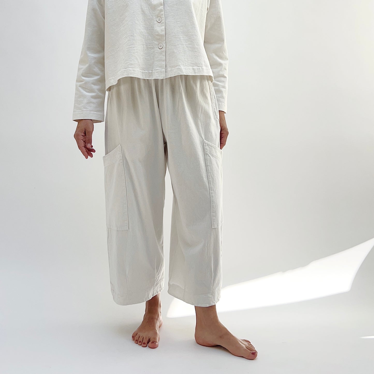 A person standing in a studio setting, wearing cream-colored cotton pants with a wide fit and high waist, paired with a matching top.