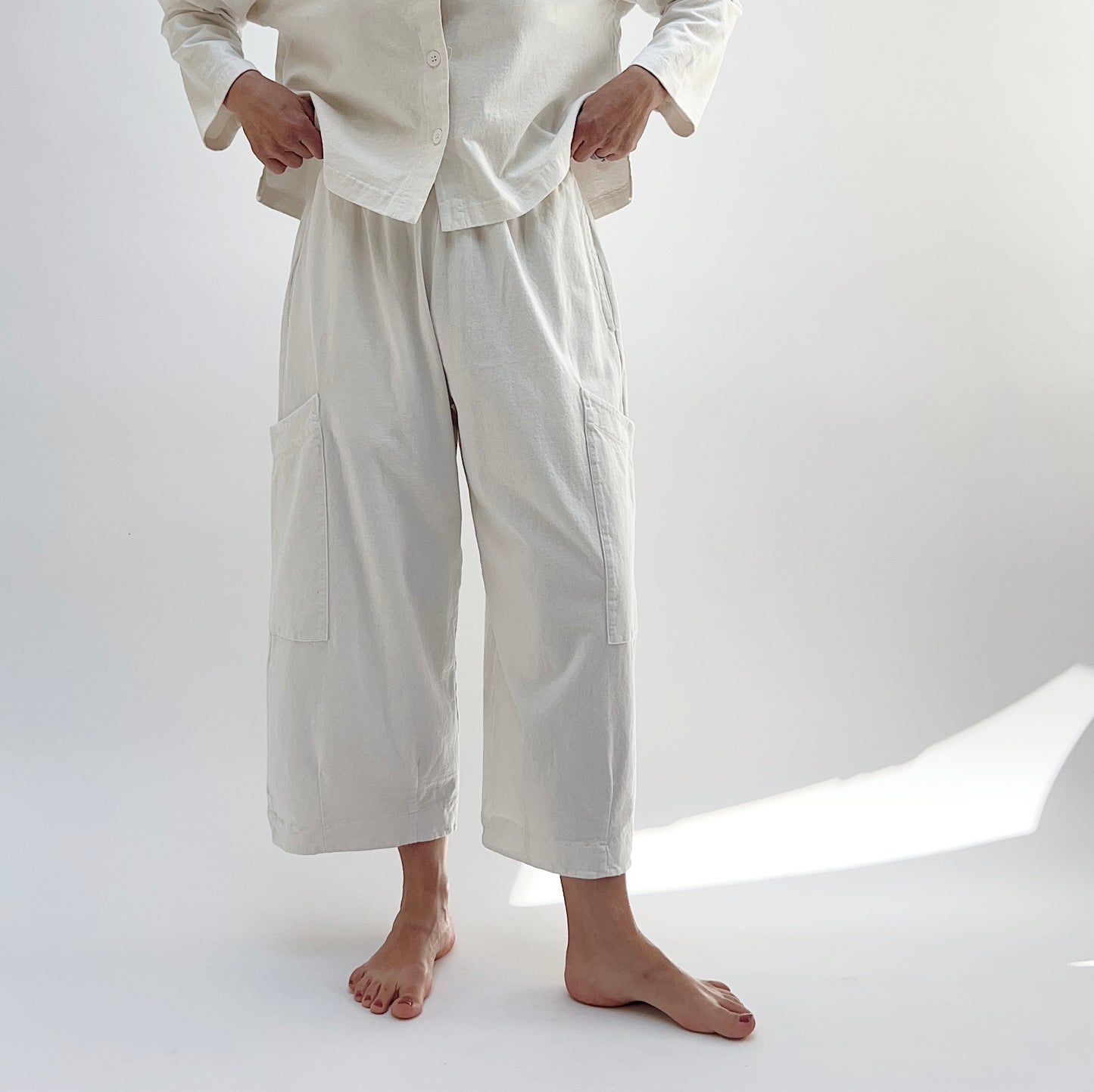A person standing in a studio setting, wearing cream-colored cotton pants with a wide fit and high waist, paired with a matching top.