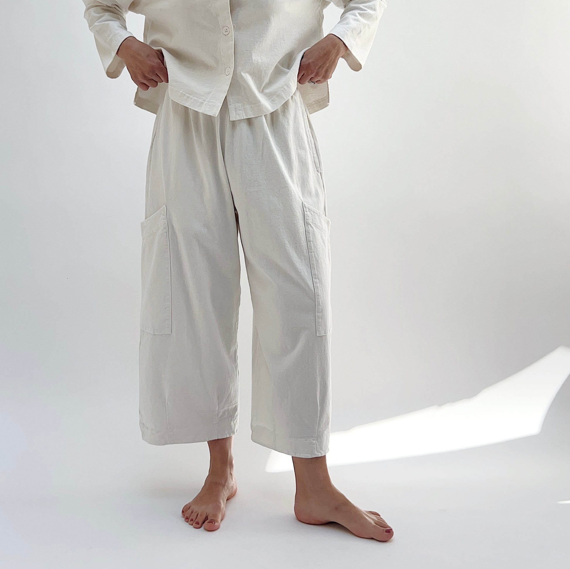 A person standing in a studio setting, wearing cream-colored cotton pants with a wide fit and high waist, paired with a matching top.