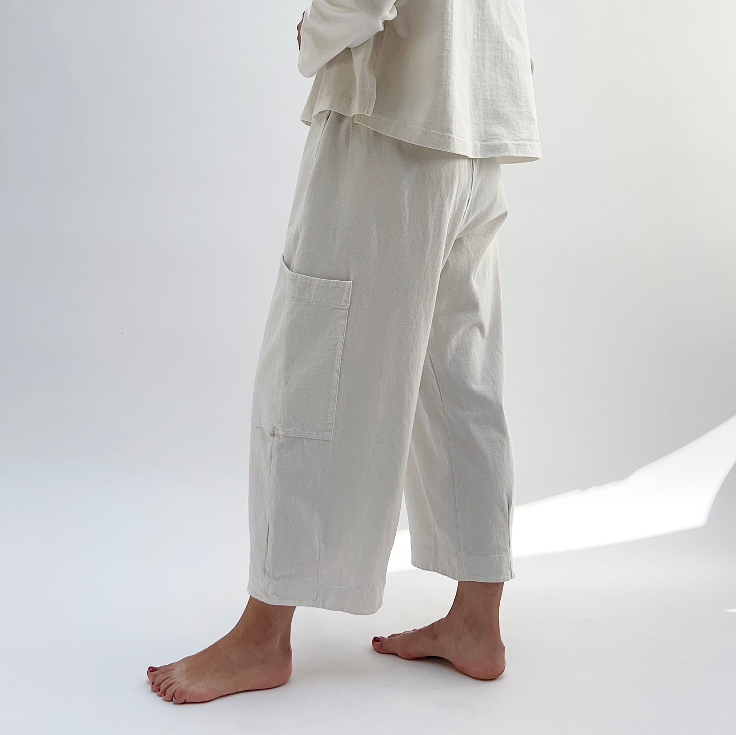 A person standing in a studio setting, wearing cream-colored cotton pants with a wide fit and high waist, paired with a matching top.