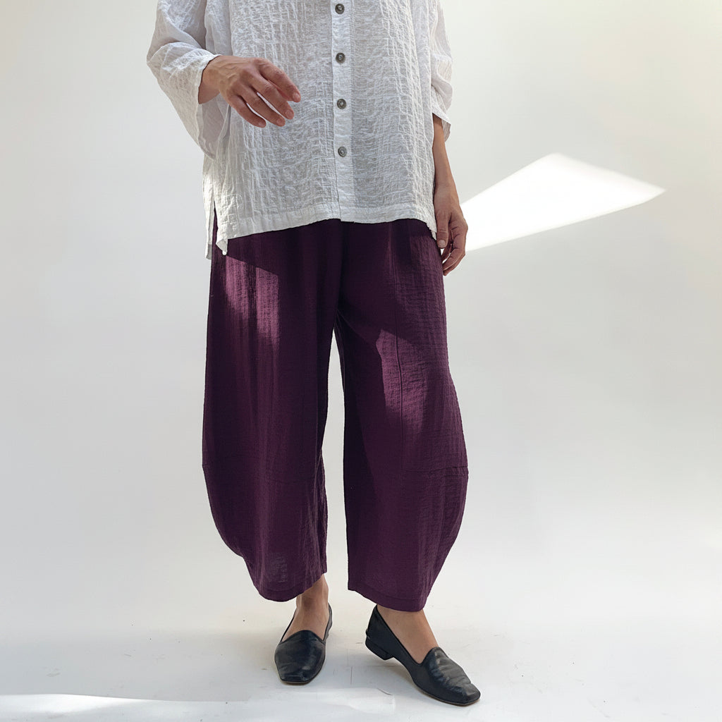 Model wearing Eleven Stitch Crinkle Travel Tucked Hem Pant in Eggplant, front view showing deep purple crinkle rayon-nylon blend, wide-leg balloon silhouette, pull-on elasticized waistband, and tucked gathered ankle hem, styled with white textured button-front top and black loafers against white studio backdrop
