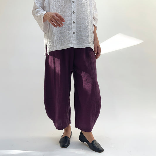 Model wearing Eleven Stitch Crinkle Travel Tucked Hem Pant in Eggplant, front view showing deep purple crinkle rayon-nylon blend, wide-leg balloon silhouette, pull-on elasticized waistband, and tucked gathered ankle hem, styled with white textured button-front top and black loafers against white studio backdrop
