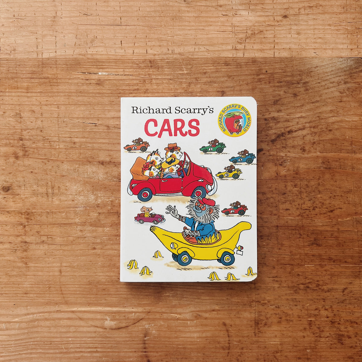 Cover of Richard Scarry's Cars board book featuring illustrated animal characters driving a red car and a yellow banana mobile, with colorful racing cars scattered across a white background
