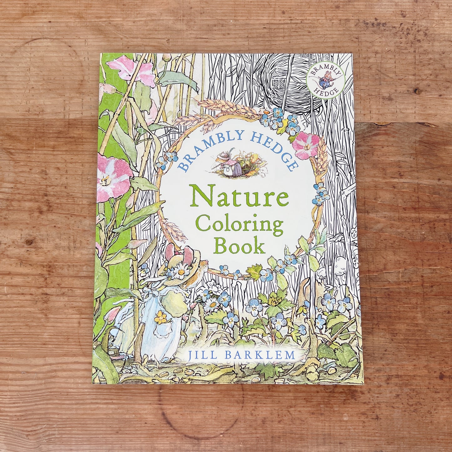 Front cover of the Brambly Hedge Nature Coloring Book by Jill Barklem, featuring a partially colored illustration of a mouse in a blue dress among pink flowers, berry-laden branches, and wheat stalks on the left, transitioning to uncolored black-and-white line art on the right, with the title in green lettering on a white oval and the Brambly Hedge series badge in the upper right corner, displayed flat on a warm wood surface.
