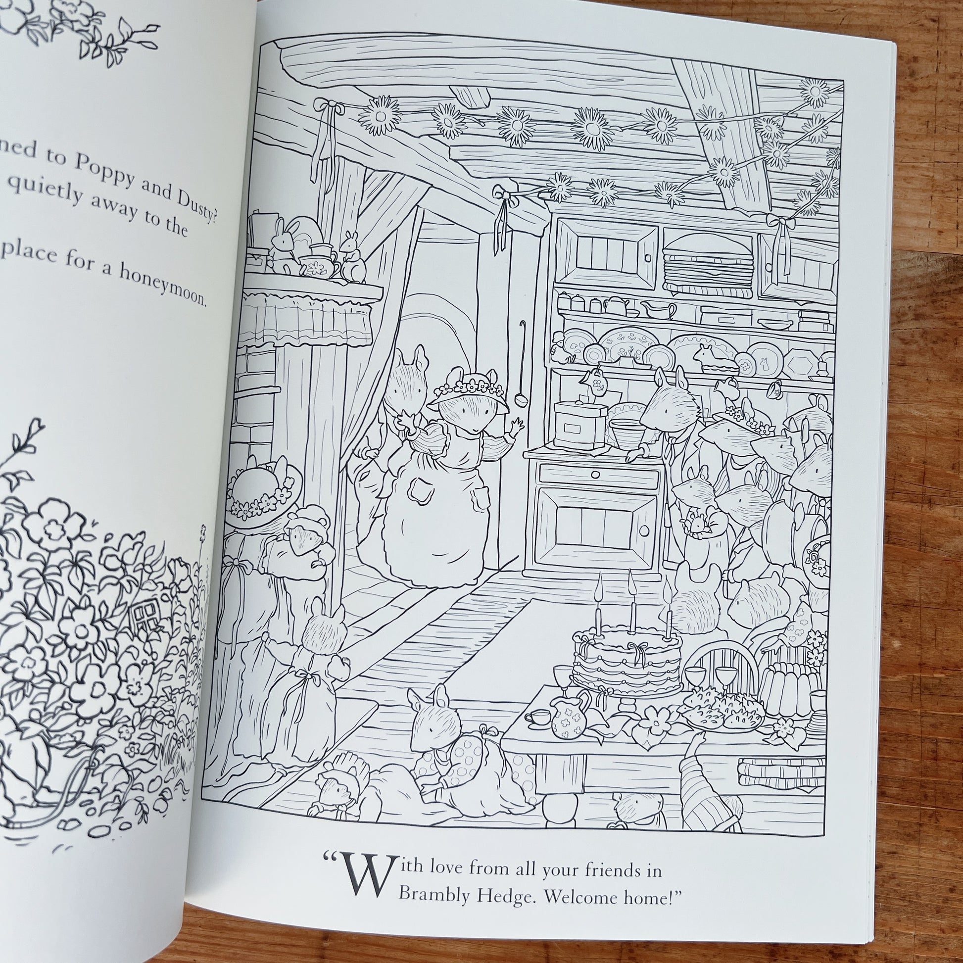Open interior spread of the Brambly Hedge Nature Coloring Book showing a full-page uncolored line-art illustration of a mouse wedding celebration in a cozy cottage kitchen: a bride mouse in a floral headpiece greets guests at the door while other mice crowd around a table set with a tiered cake, teacups, and food, with shelves of crockery, hanging flower garlands, and a quote reading "With love from all your friends in Brambly Hedge. Welcome home!" below, displayed on a warm wood surface.
