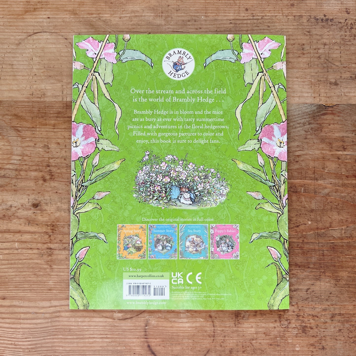 Back cover of the Brambly Hedge Nature Coloring Book with a green background bordered by pink sweet pea flowers and stems, featuring the Brambly Hedge circular logo at top, a descriptive blurb, a central illustration of mice gathered outside a hedgerow cottage, thumbnail covers of the four original stories Spring Story, Summer Story, Sea Story, and Poppy's Babies, and a barcode with retail price of $10.99 and "Suitable for ages 3+" notation, displayed flat on a warm wood surface.
