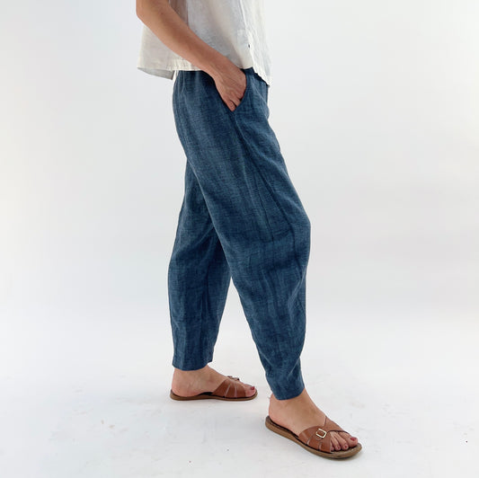 Person wearing blue melange linen pants and a white top and brown sandals, with their hands in their pockets on a white background.