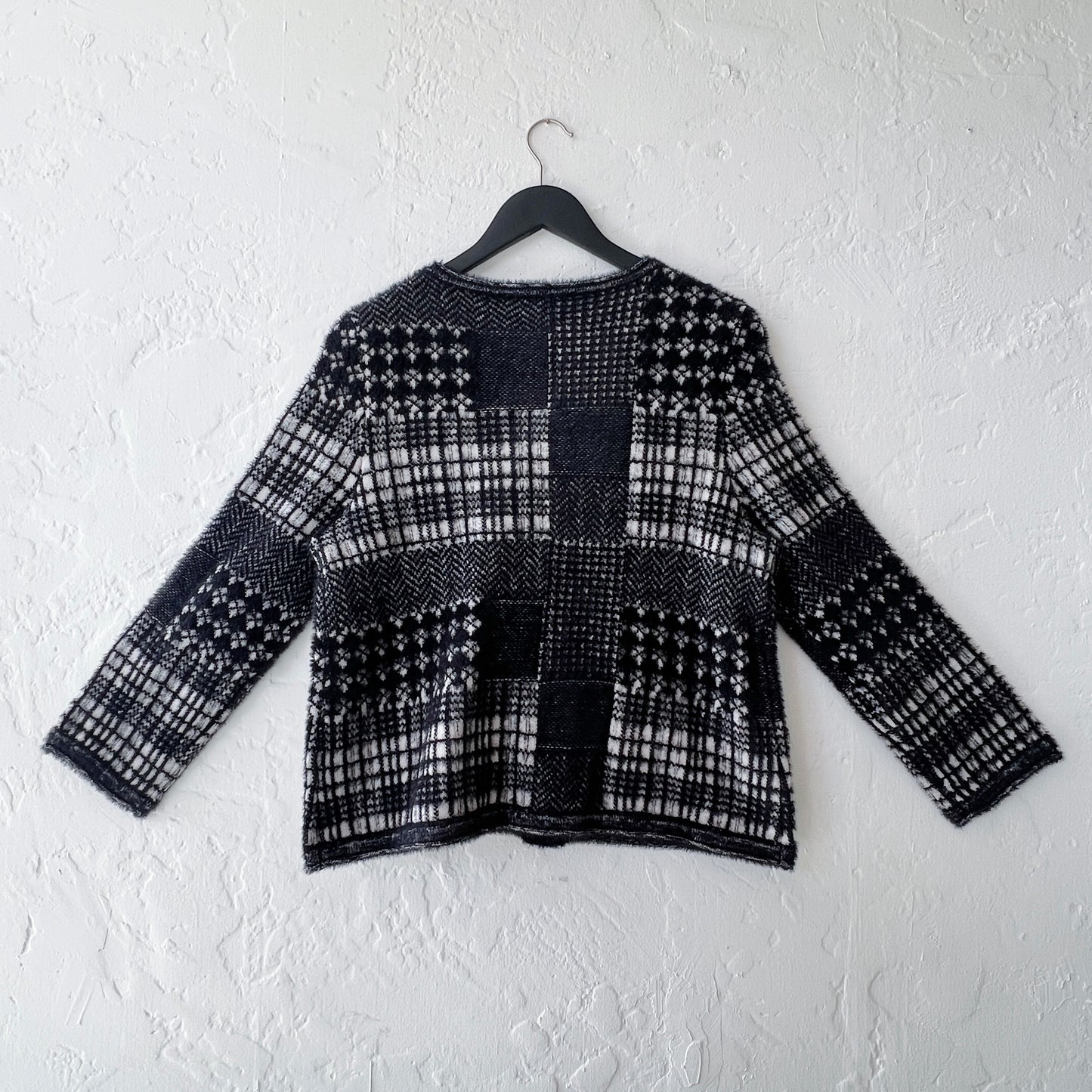 Habitat Plaid Patchwork Cardigan on hanger, back view showing mixed Fair Isle, houndstooth, herringbone, and windowpane plaid knit panels in black and cream with soft eyelash texture, long sleeves with ribbed cuffs, and relaxed boxy silhouette
