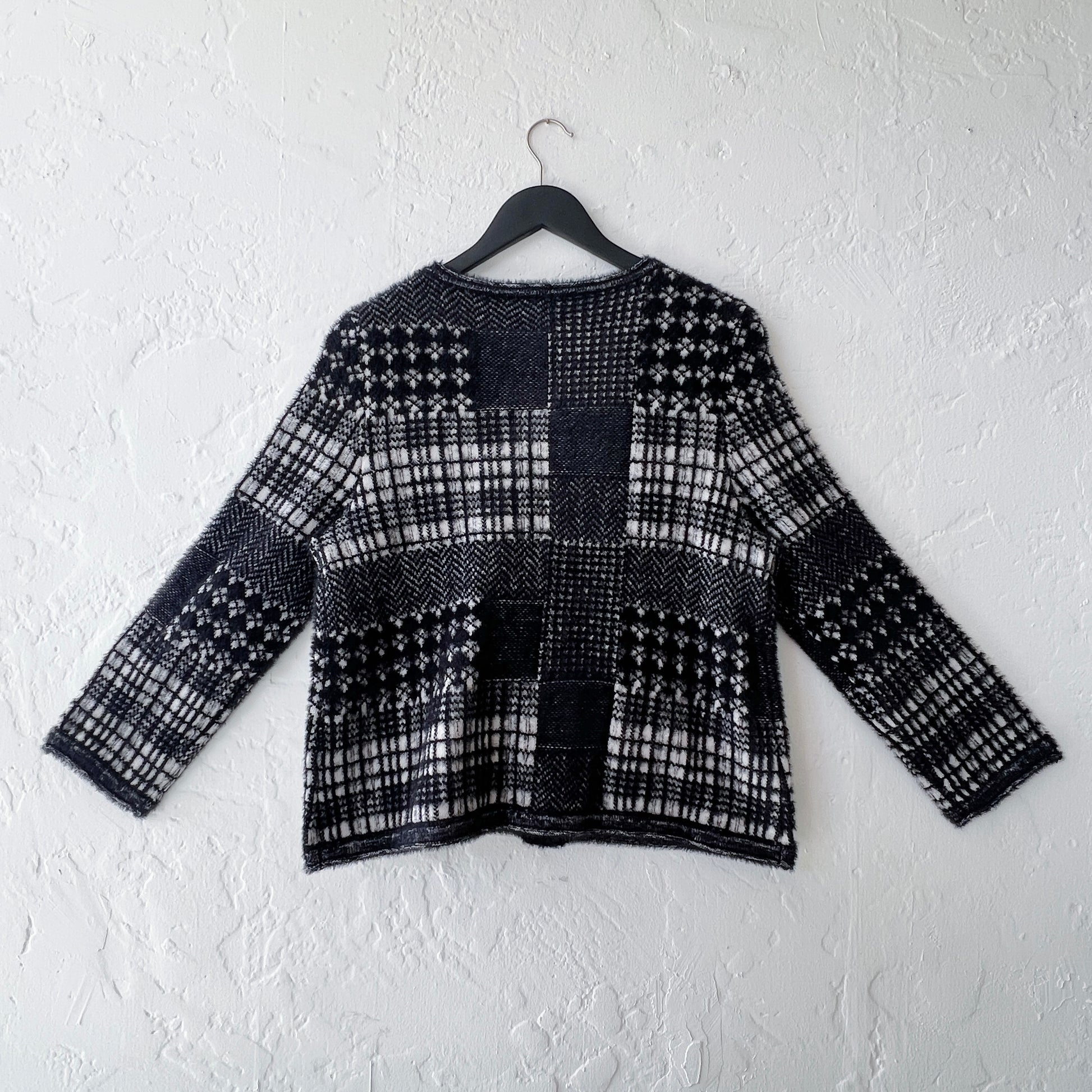 Habitat Plaid Patchwork Cardigan on hanger, back view showing mixed Fair Isle, houndstooth, herringbone, and windowpane plaid knit panels in black and cream with soft eyelash texture, long sleeves with ribbed cuffs, and relaxed boxy silhouette
