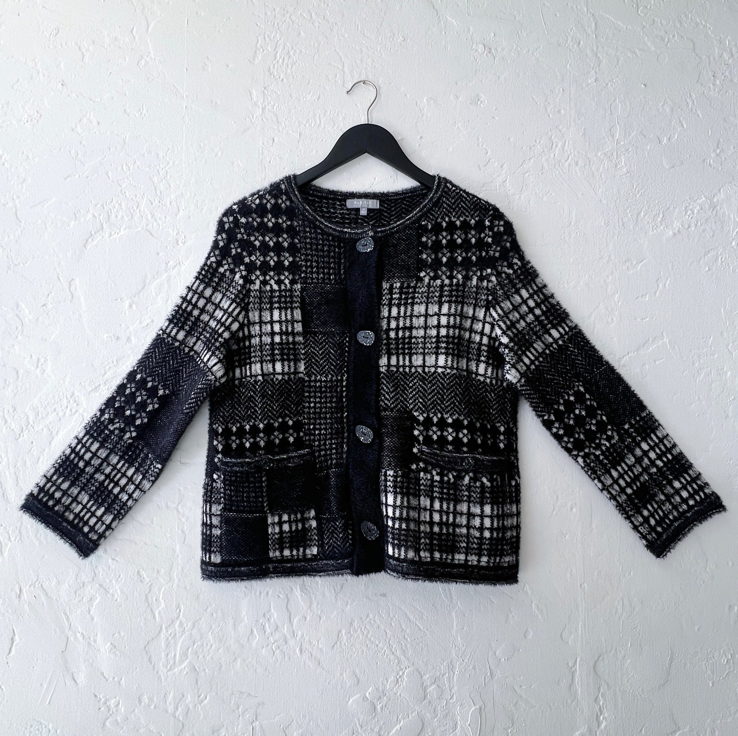 Habitat Plaid Patchwork Cardigan on hanger, front view showing mixed Fair Isle, houndstooth, herringbone, and windowpane plaid knit panels in black and cream with soft eyelash texture, collarless crewneck, 4-button placket with speckled resin buttons, and two button-flap hip pockets
