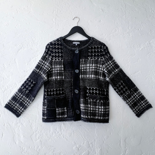 Habitat Plaid Patchwork Cardigan on hanger, front view showing mixed Fair Isle, houndstooth, herringbone, and windowpane plaid knit panels in black and cream with soft eyelash texture, collarless crewneck, 4-button placket with speckled resin buttons, and two button-flap hip pockets
