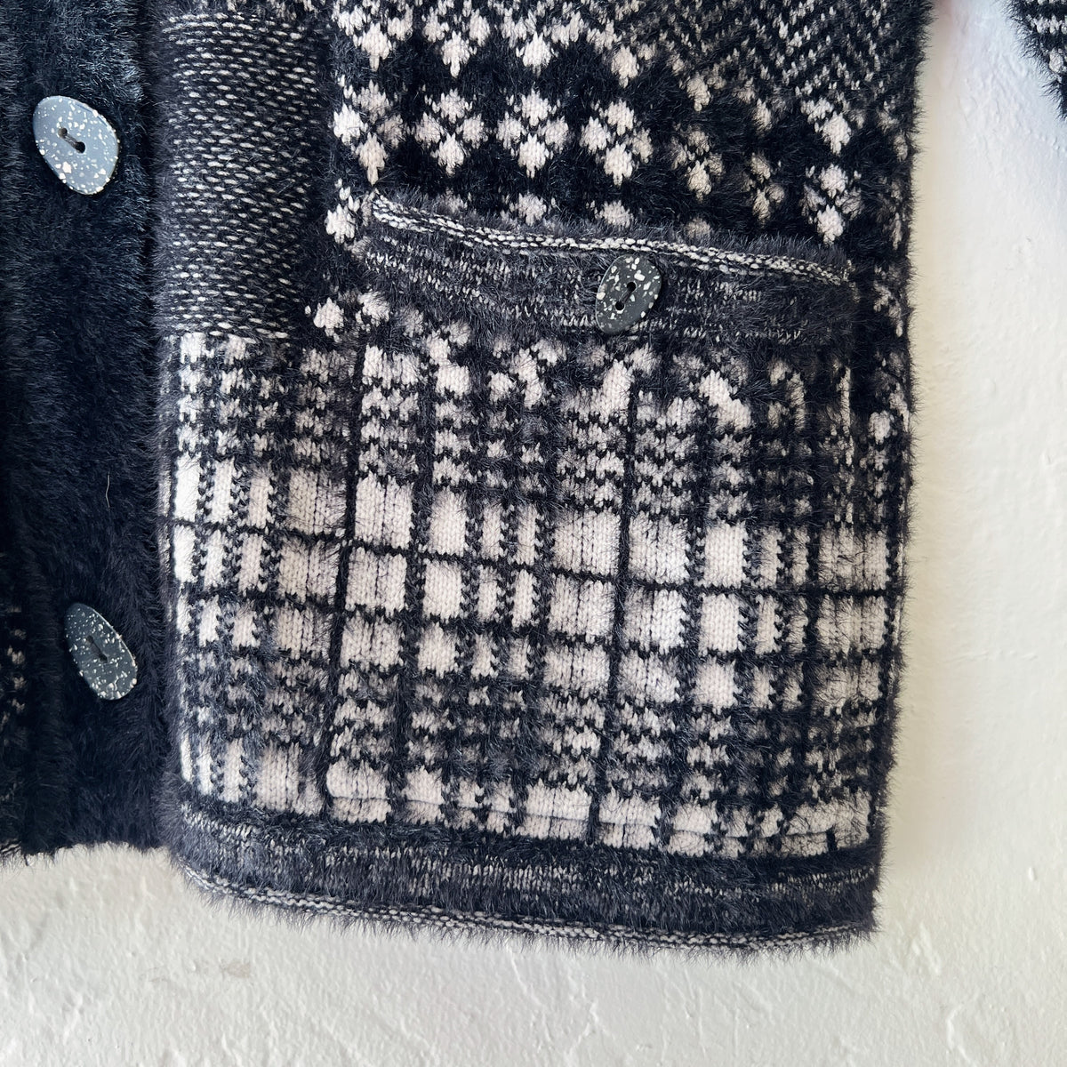 Close-up detail of Habitat Plaid Patchwork Cardigan showing button-flap hip pocket with speckled resin button, mixed plaid and Fair Isle knit panels in black and cream, and soft eyelash-textured nylon blend fabric
