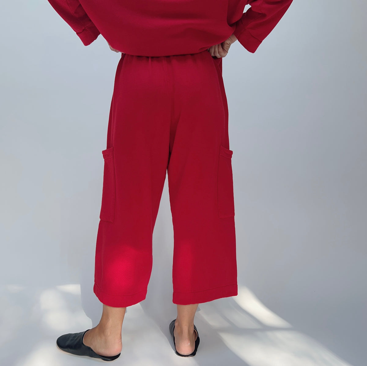 Back view of model wearing the Bryn Walker Pasha Fleece Pant in Jester, showing elastic waistband, two large outer thigh patch pockets, wide cropped leg, mid-calf length, and vivid jester red organic cotton-bamboo fleece, styled with matching Pasha Fleece Top and black leather mules
