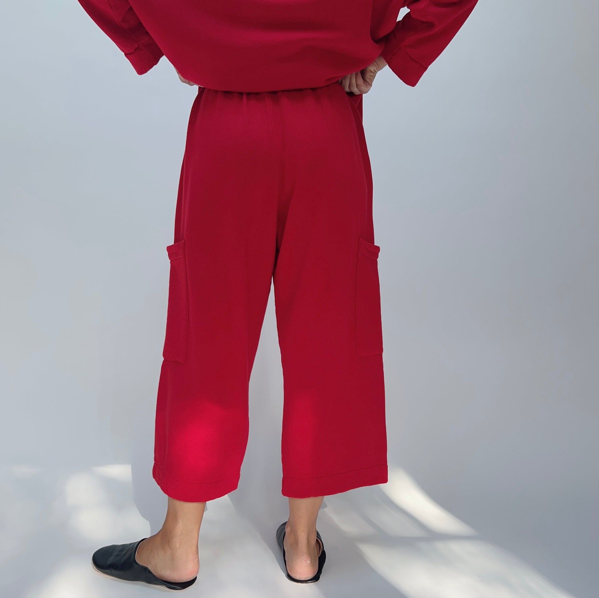 Back view of model wearing the Bryn Walker Pasha Fleece Pant in Jester, showing elastic waistband, two large outer thigh patch pockets, wide cropped leg, mid-calf length, and vivid jester red organic cotton-bamboo fleece, styled with matching Pasha Fleece Top and black leather mules
