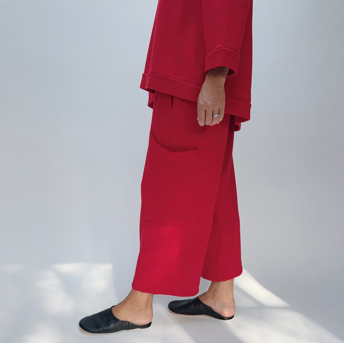 Side view of model wearing the Bryn Walker Pasha Fleece Pant in Jester, showing elastic waistband, large side pocket, wide cropped leg silhouette, mid-calf length, and vivid jester red organic cotton-bamboo fleece, styled with matching Pasha Fleece Top and black leather mules
