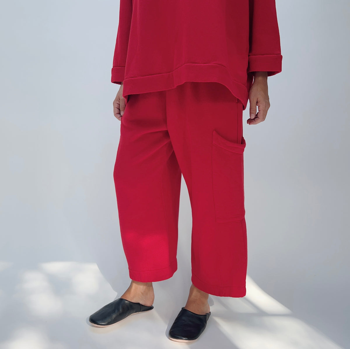 Model wearing the Bryn Walker Pasha Fleece Pant in Jester, angled front view showing elastic waistband, large outer thigh patch pocket, wide cropped leg, mid-calf length, and vivid jester red organic cotton-bamboo fleece, styled with matching Pasha Fleece Top and black leather mules
