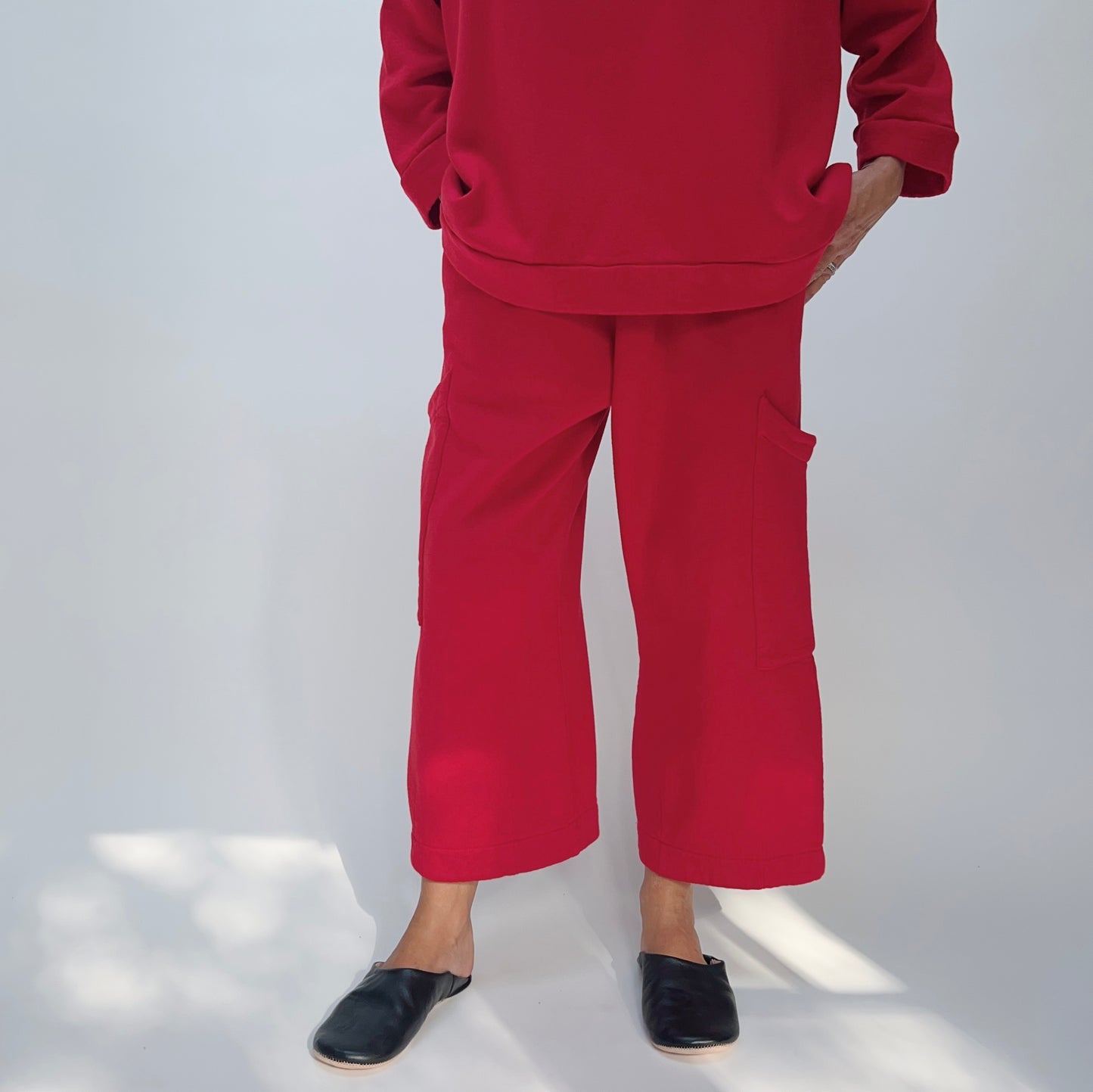 Model with hands in pockets wearing the Bryn Walker Pasha Fleece Pant in Jester, full front view showing elastic waistband, two large outer thigh patch pockets, wide cropped leg, mid-calf length, and vivid jester red organic cotton-bamboo fleece, styled with matching Pasha Fleece Top and black leather mules

