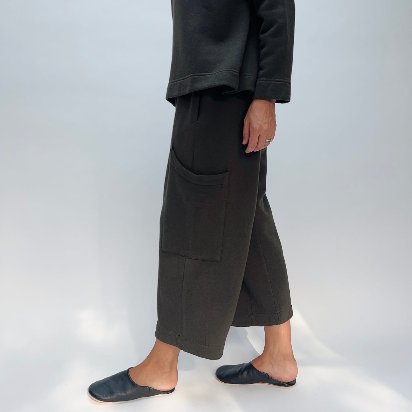 Side view of model wearing the Bryn Walker Pasha Fleece Pant in Kelp, showing elastic waistband, large outer thigh patch pocket, wide cropped leg silhouette, folded cuff hem, and deep forest green organic cotton-bamboo fleece, styled with matching Pasha Fleece Top and black leather mules
