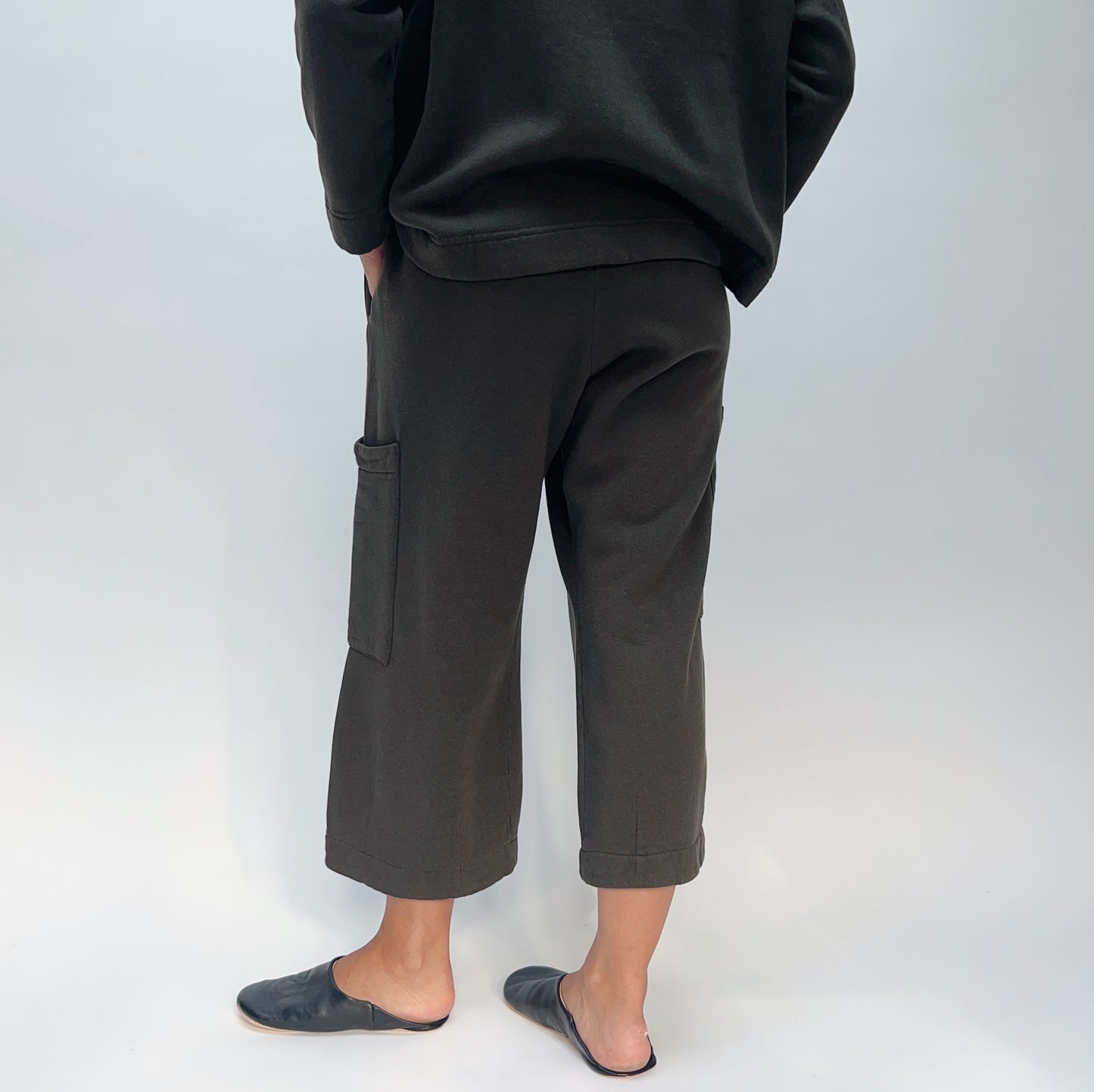Back view of model wearing the Bryn Walker Pasha Fleece Pant in Kelp, showing elastic waistband, two large outer thigh patch pockets, wide cropped leg, folded cuff hem, and deep forest green organic cotton-bamboo fleece, styled with matching Pasha Fleece Top and black leather mules

