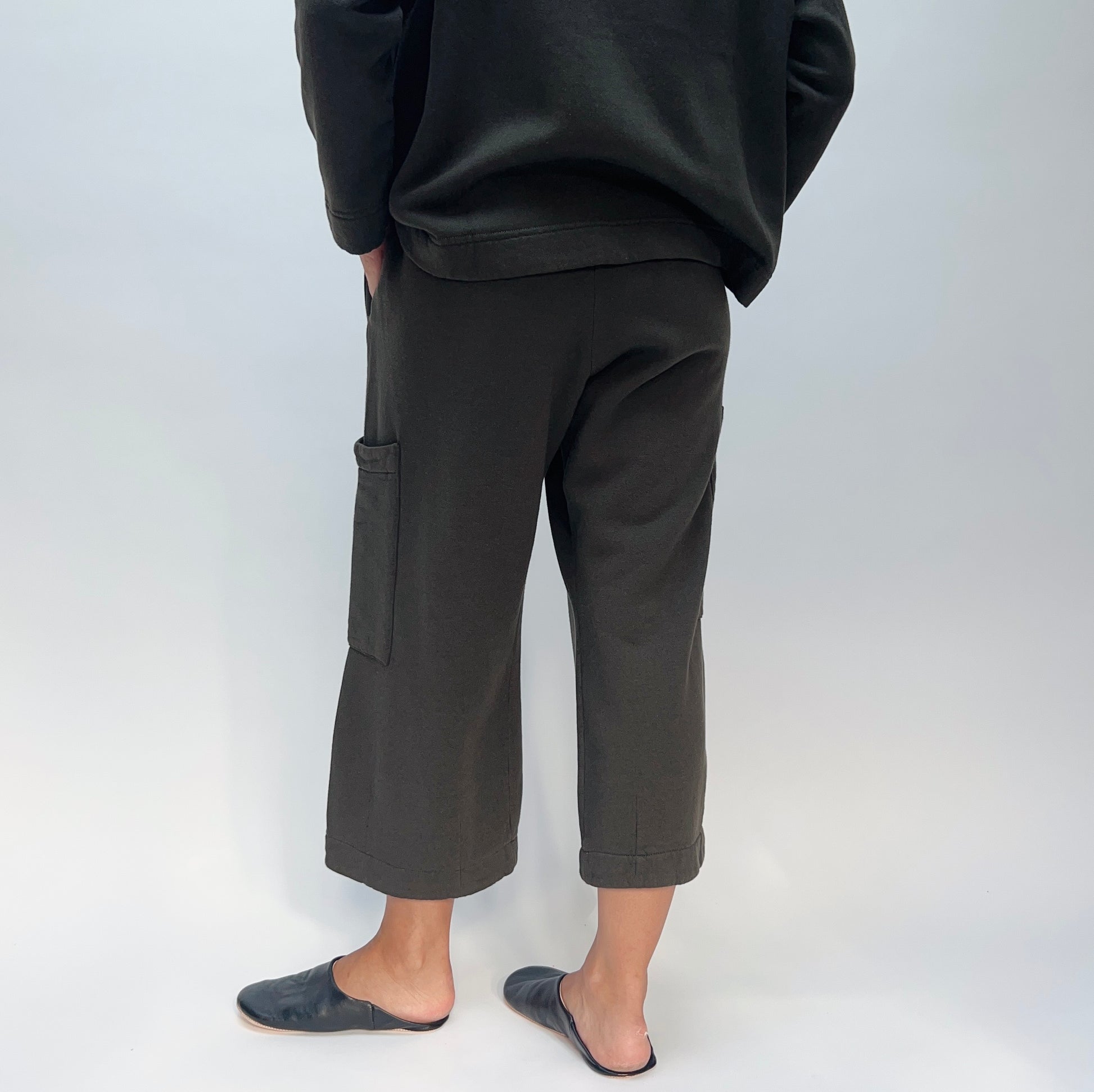 Back view of model wearing the Bryn Walker Pasha Fleece Pant in Kelp, showing elastic waistband, two large outer thigh patch pockets, wide cropped leg, folded cuff hem, and deep forest green organic cotton-bamboo fleece, styled with matching Pasha Fleece Top and black leather mules
