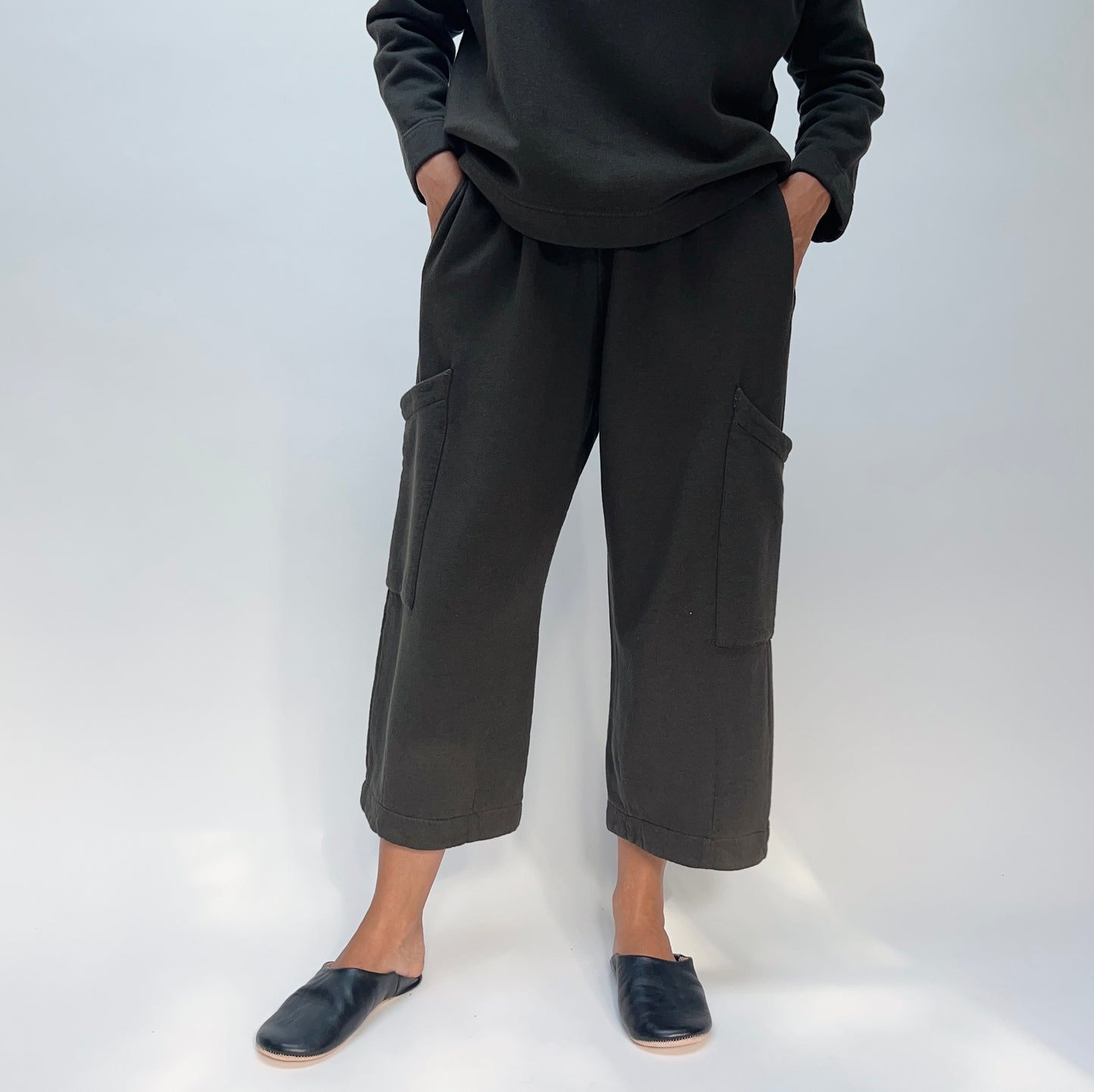 Model with hands in pockets wearing the Bryn Walker Pasha Fleece Pant in Kelp, full front view showing elastic waistband, two large outer thigh patch pockets, wide cropped leg, folded cuff hem, and deep forest green organic cotton-bamboo fleece, styled with matching Pasha Fleece Top and black leather mules
