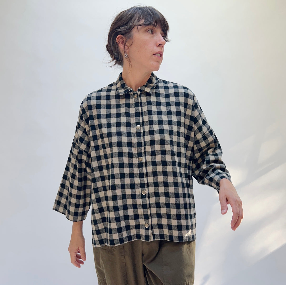 Model wearing the Eleven Stitch Big Check Big Shirt in Fennel, three-quarter front view, boxy snap-button shirt in black and tan gingham cotton, paired with olive trousers
