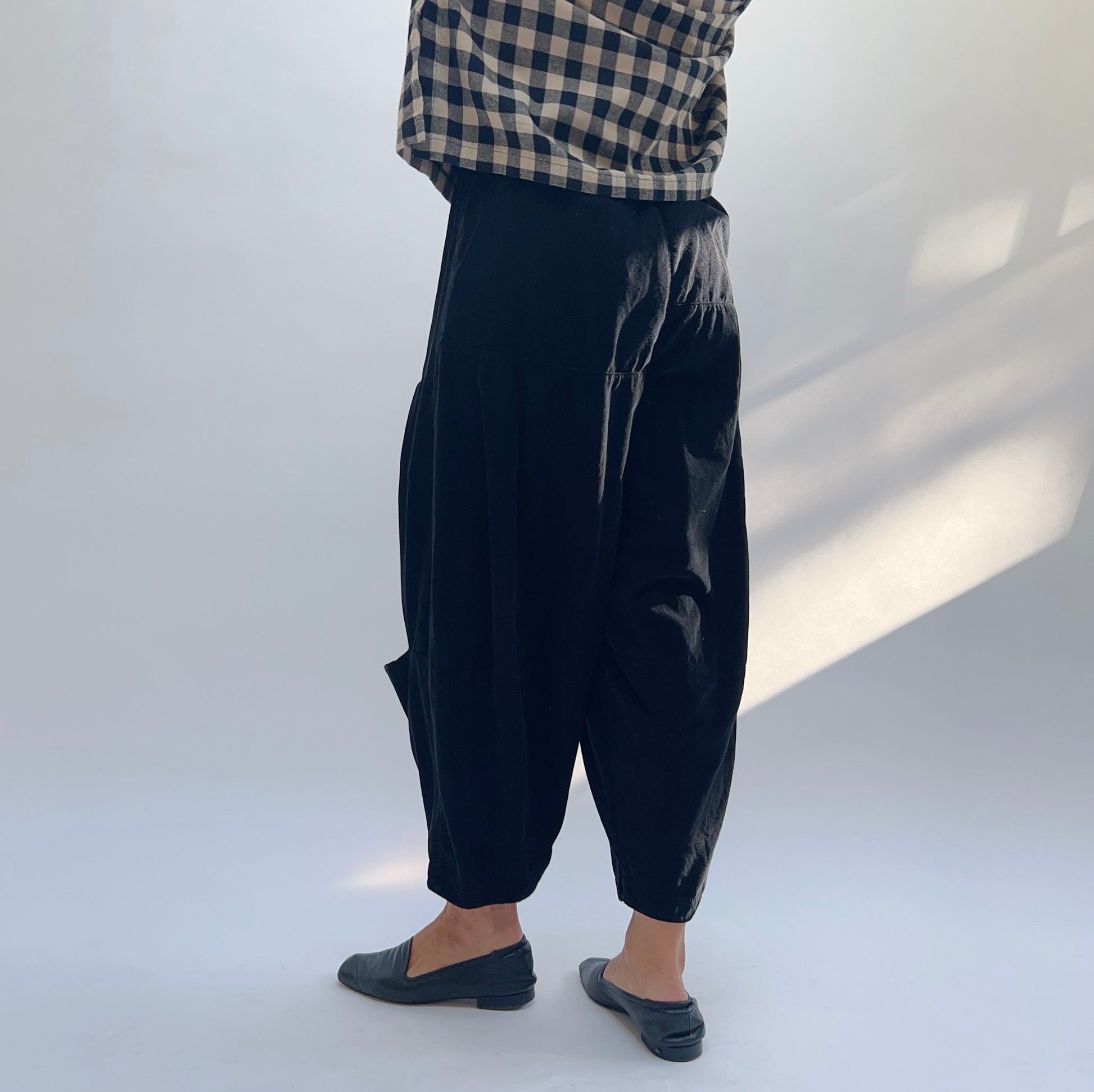 Side view of black corduroy pant showing elasticated waistband, voluminous drape, and tapered ankle with side pocket
