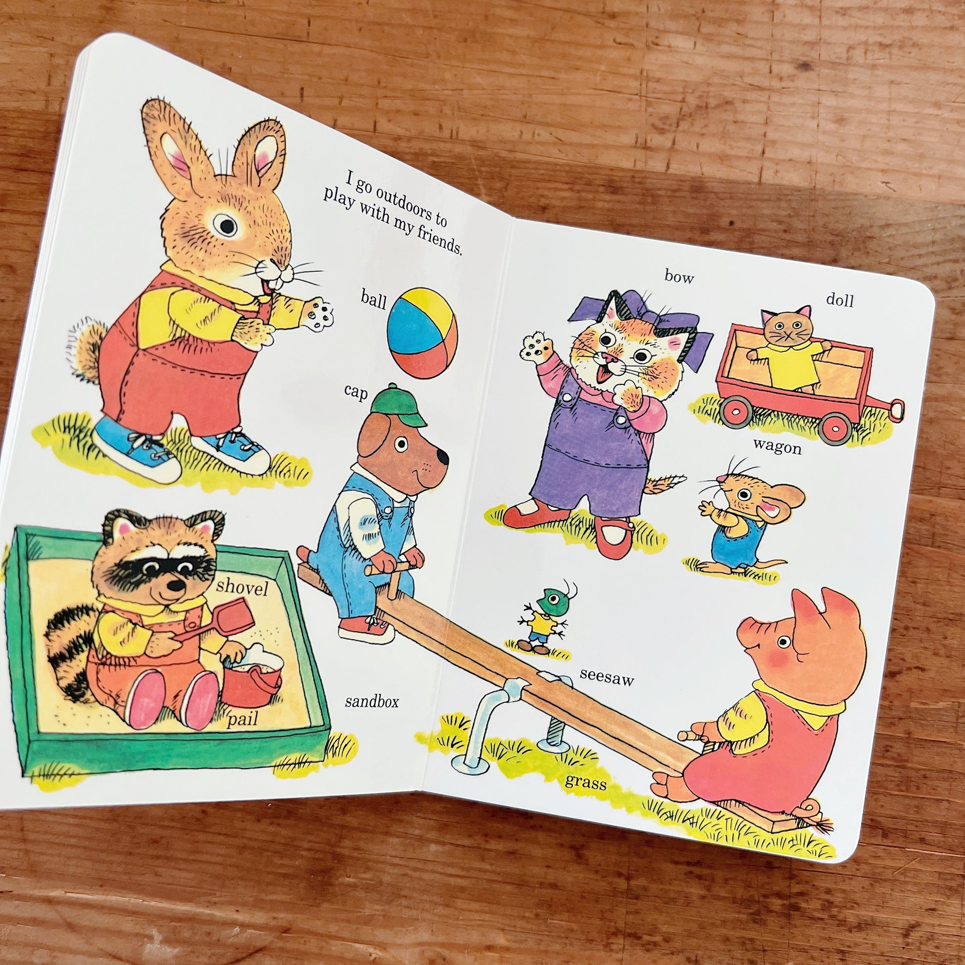Open interior spread of Richard Scarry's Best Little Board Book Ever showing the playground page from the Early Words section, with white background and Richard Scarry's color illustrations of animal characters at play. Header text reads "I go outdoors to play with my friends." Labeled objects include: Frannie the bunny (upper left), a raccoon in a sandbox with a shovel and pail, a dog in a cap, a colorful ball, a cat with a bow in a purple dress waving, a small mouse in blue overalls, a pig on a seesaw, a 
