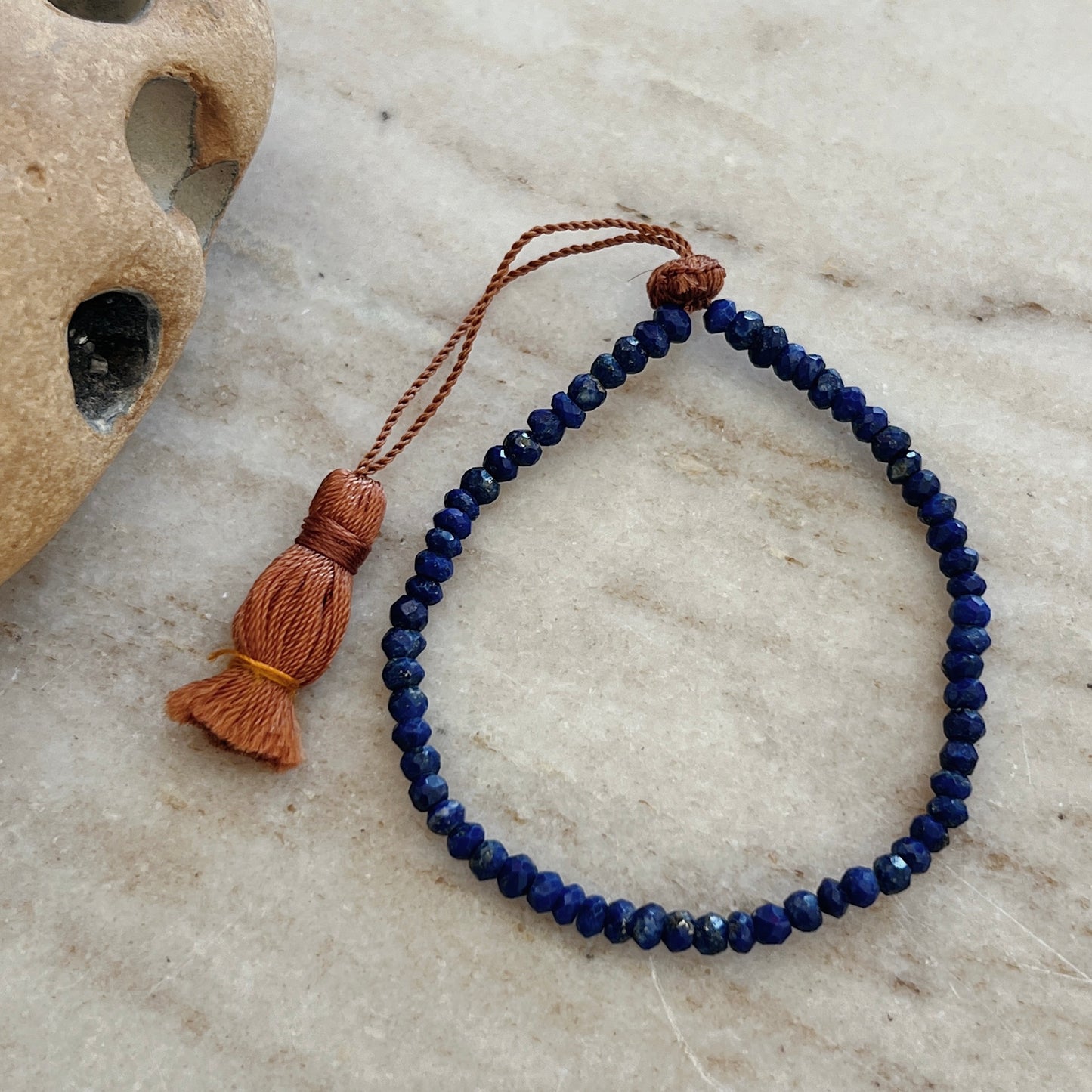 A faceted lapis bead bracelet with an adjustable tassel closure, displayed on a textured surface.