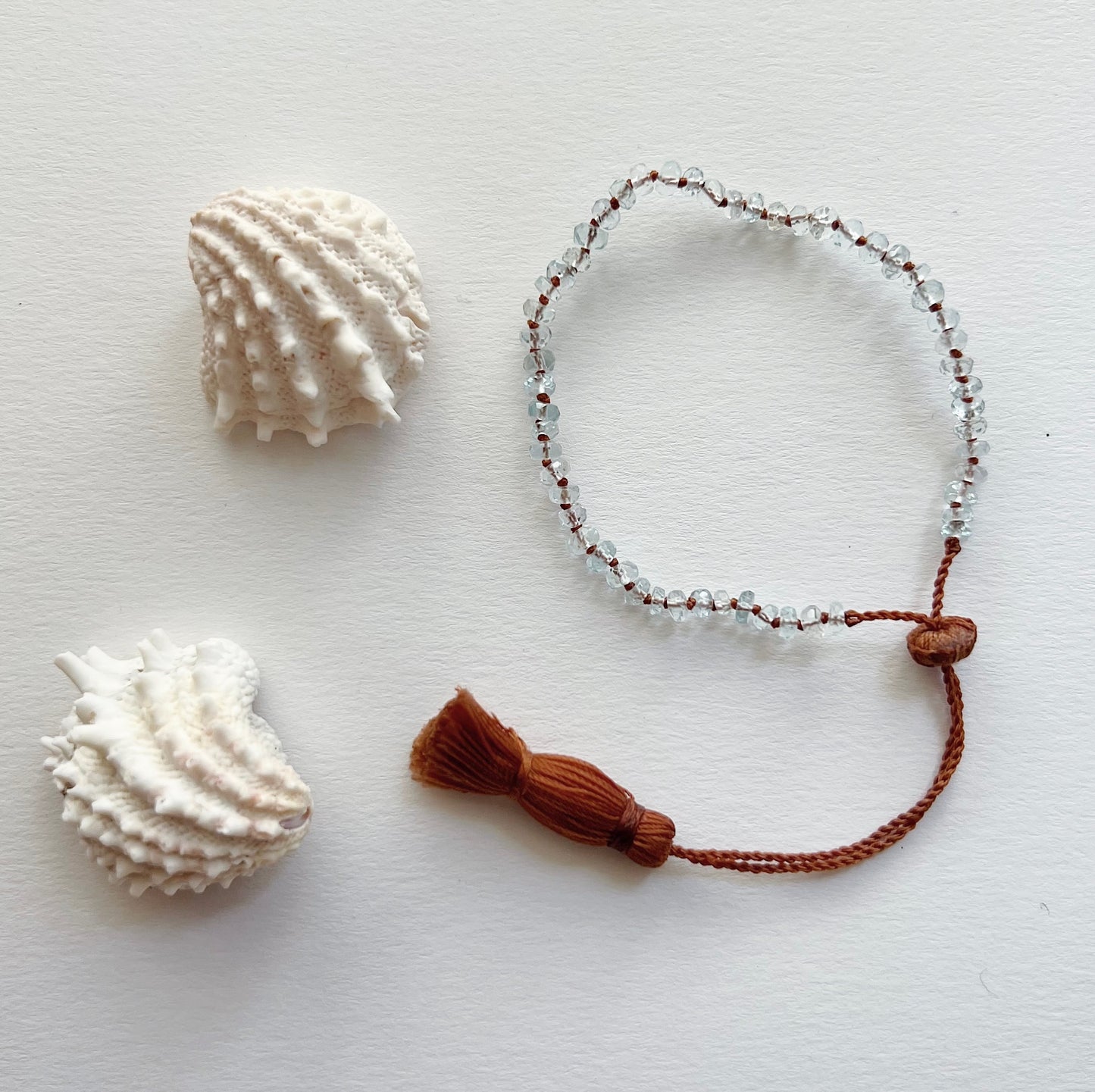 An aquamarine beaded bracelet with a waxed silk cord and an adjustable tassel closure, displayed next to two seashells.