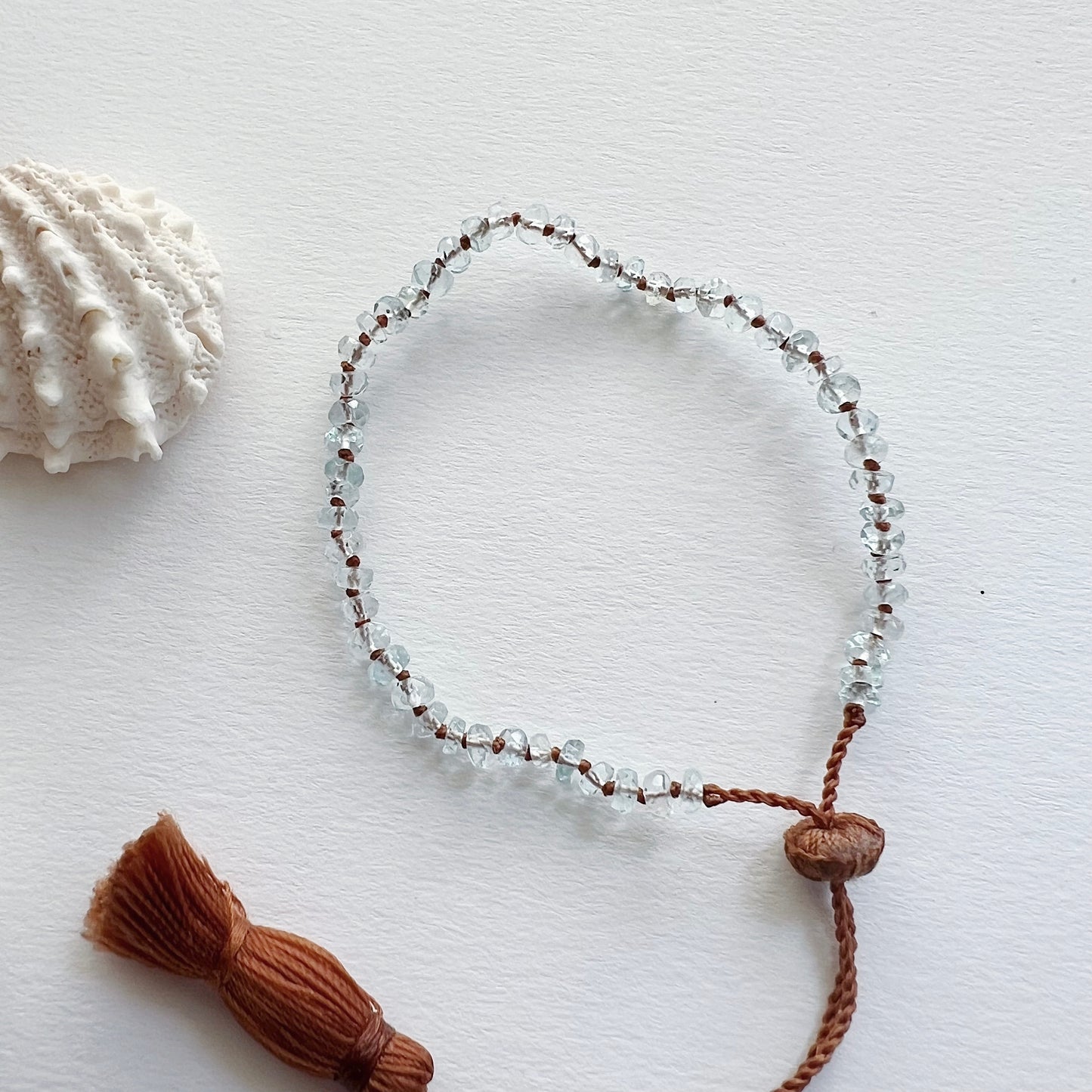 An aquamarine beaded bracelet with a waxed silk cord and an adjustable tassel closure, displayed next to a seashell.