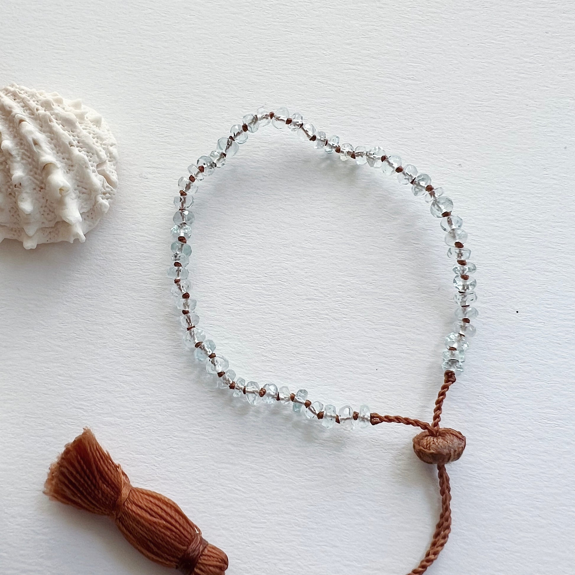 An aquamarine beaded bracelet with a waxed silk cord and an adjustable tassel closure, displayed next to a seashell.