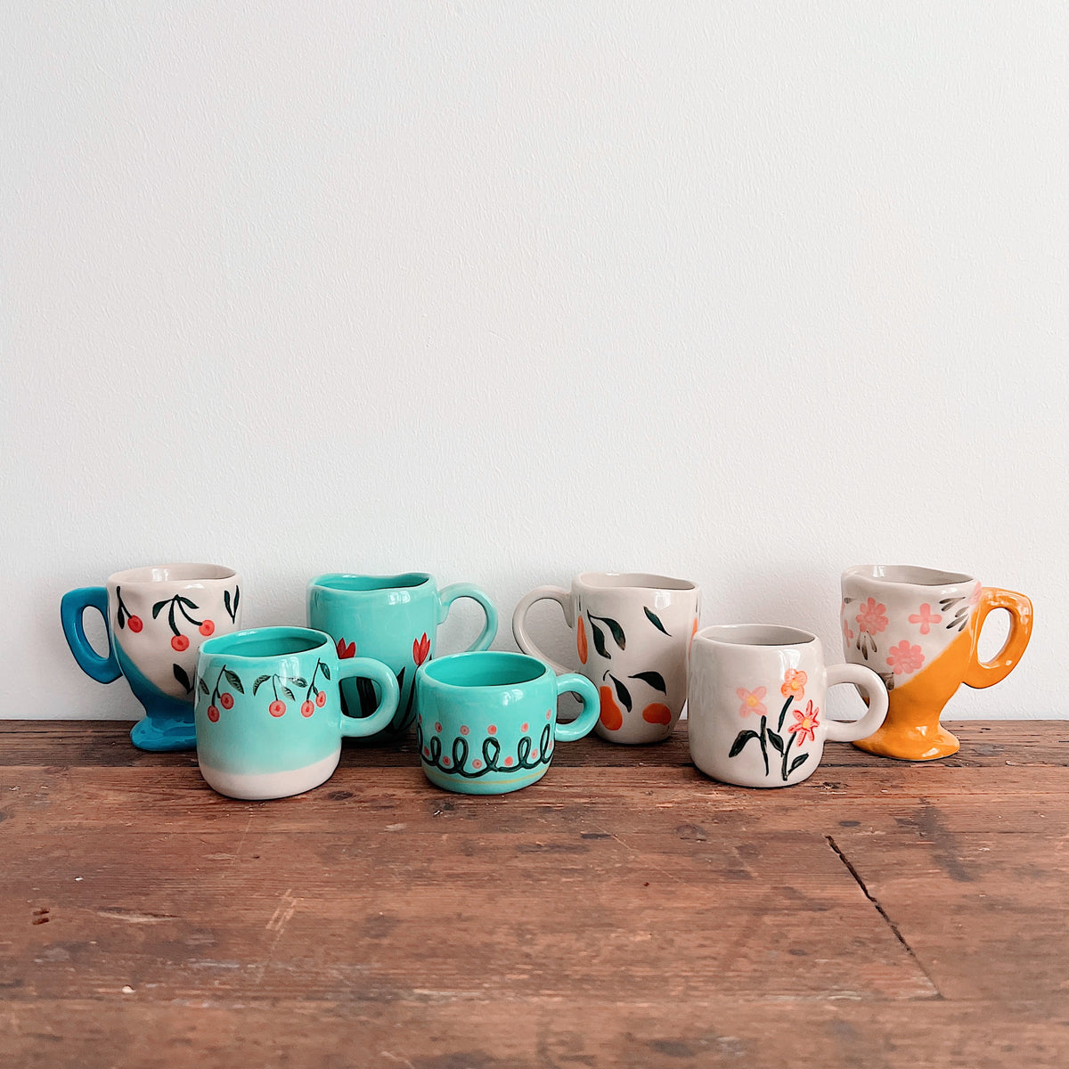 Seven assorted hand-painted stoneware mugs with cherry, fruit, floral, and looping patterns, arranged on a wooden tabletop.
