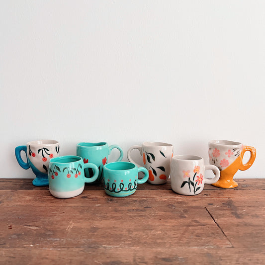 Seven assorted hand-painted stoneware mugs with cherry, fruit, floral, and looping patterns, arranged on a wooden tabletop.
