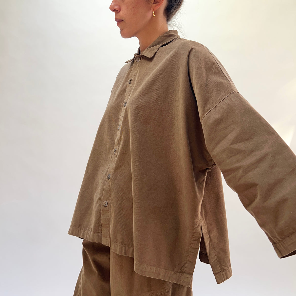 Three-quarter side view of a woman wearing the Eleven Stitch Corduroy Big Shirt in Winter Olive with arm extended, showing the point collar, shell button front, dropped shoulder seam, wide sleeve with banded cuff, side vent at the hem, and the boxy oversized silhouette in fine-wale olive cotton corduroy, styled with matching corduroy wide-leg trousers against a white background
