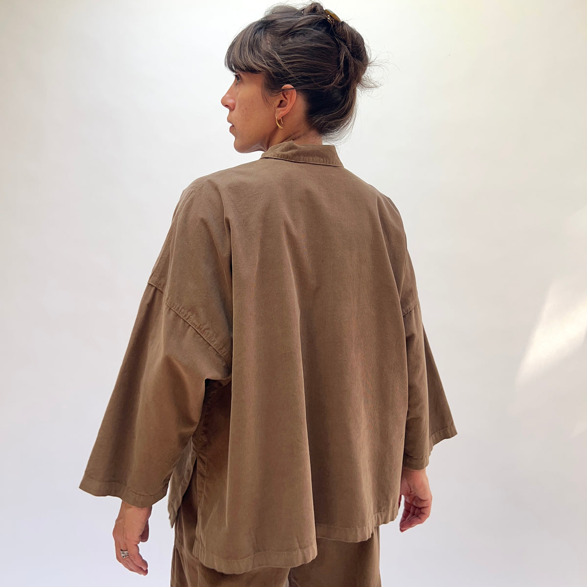 Back view of a woman wearing the Eleven Stitch Corduroy Big Shirt in Winter Olive, showing the dropped shoulder seam with topstitching, wide sleeves, straight cropped back hem with side vents, and the boxy oversized silhouette in fine-wale olive cotton corduroy, styled with matching corduroy wide-leg trousers against a white background
