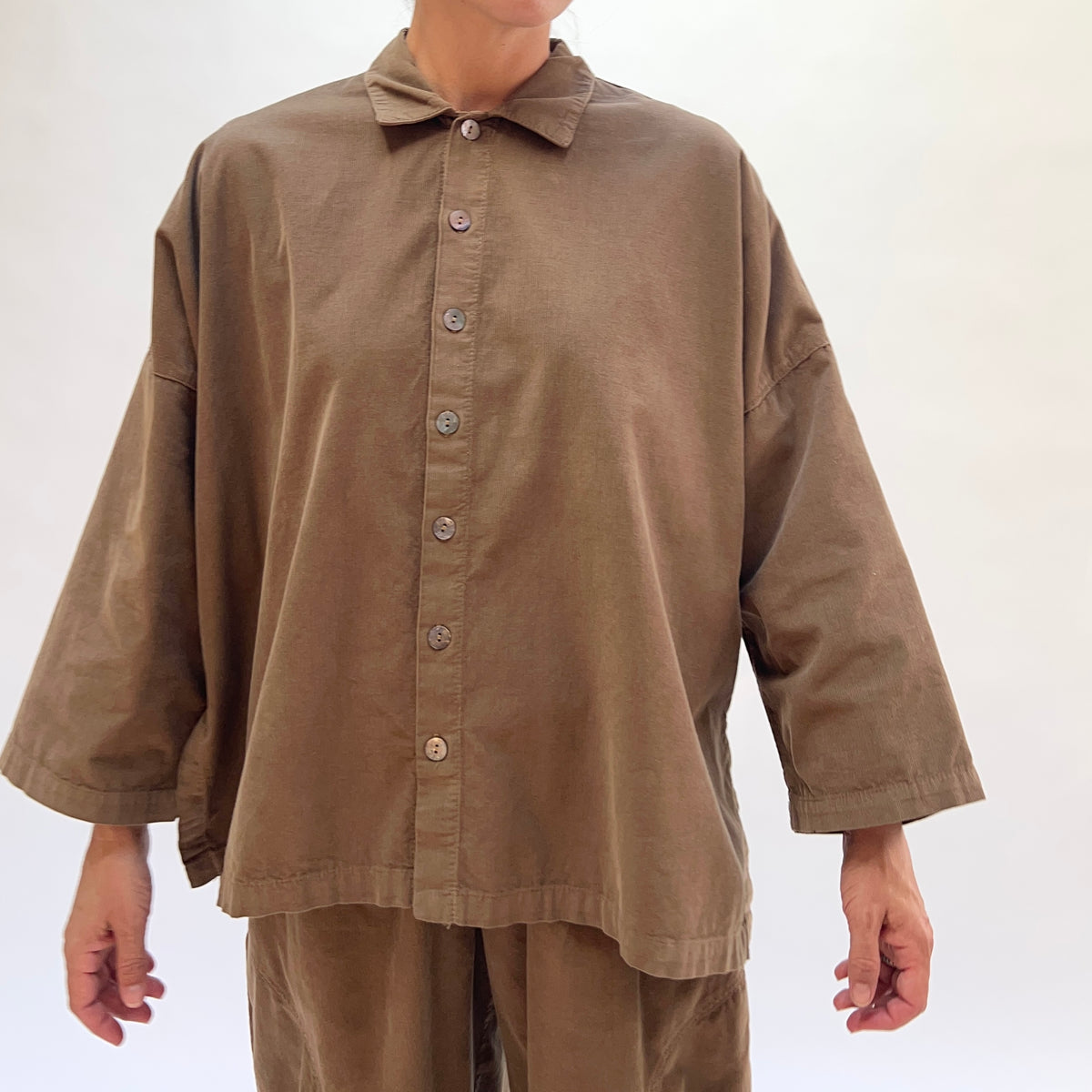 Front view of the Eleven Stitch Corduroy Big Shirt in Winter Olive on a model, showing the classic point collar, full shell button front placket, dropped shoulder seams with topstitching, wide sleeves with banded cuffs, straight cropped hem, and the boxy oversized silhouette in fine-wale olive cotton corduroy, styled with matching corduroy wide-leg trousers against a white background
