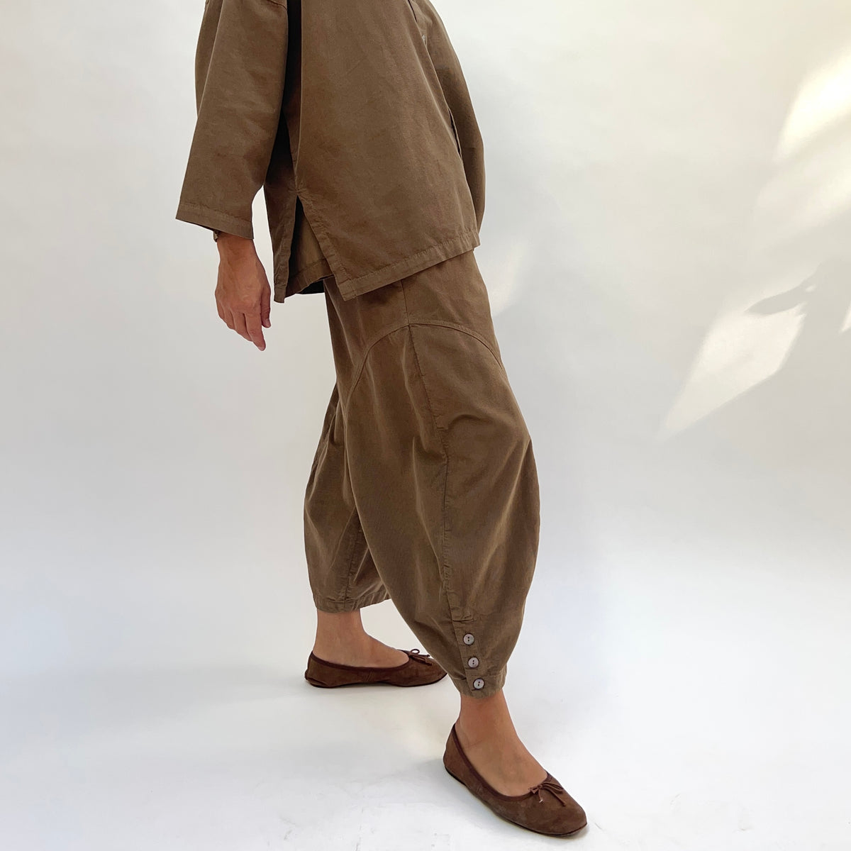 Side view of a woman wearing the Eleven Stitch Corduroy Doreen Pant in Winter Olive, showing the curved panel seam on the thigh, relaxed balloon leg, gathered ankle with three shell buttons at the cuff, in fine-wale olive cotton corduroy, styled with the matching corduroy shirt and brown ballet flats against a white background

