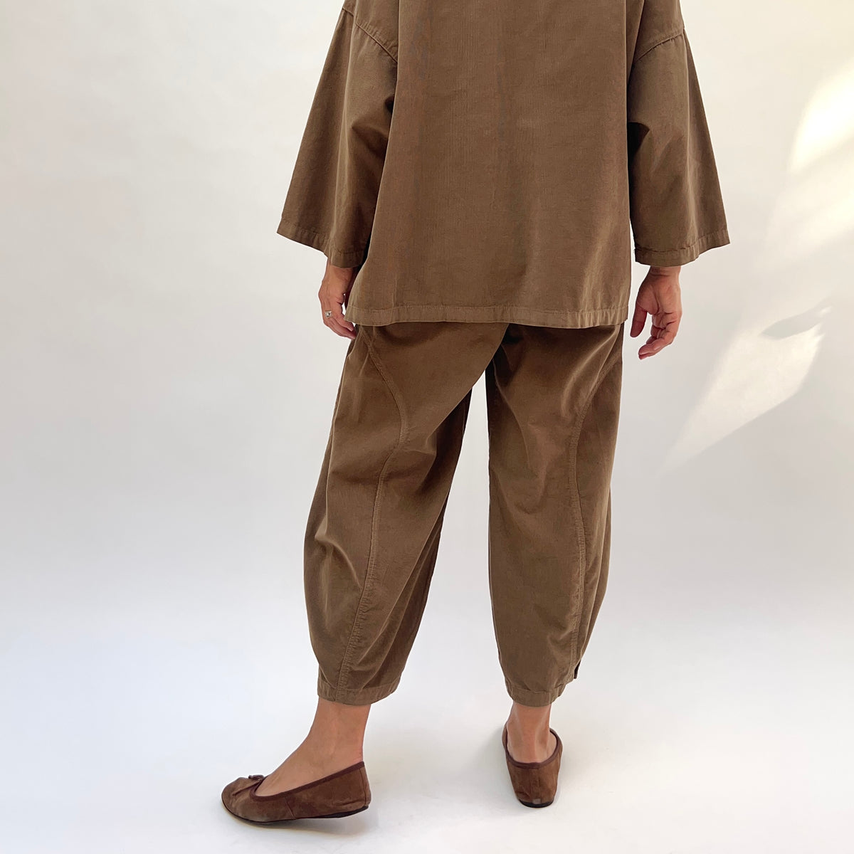 Back view of a woman wearing the Eleven Stitch Corduroy Doreen Pant in Winter Olive, showing the relaxed high-rise back, curved balloon leg tapering to a gathered ankle, three shell buttons at the cuff, in fine-wale olive cotton corduroy, styled with the matching corduroy shirt and brown ballet flats against a white background
